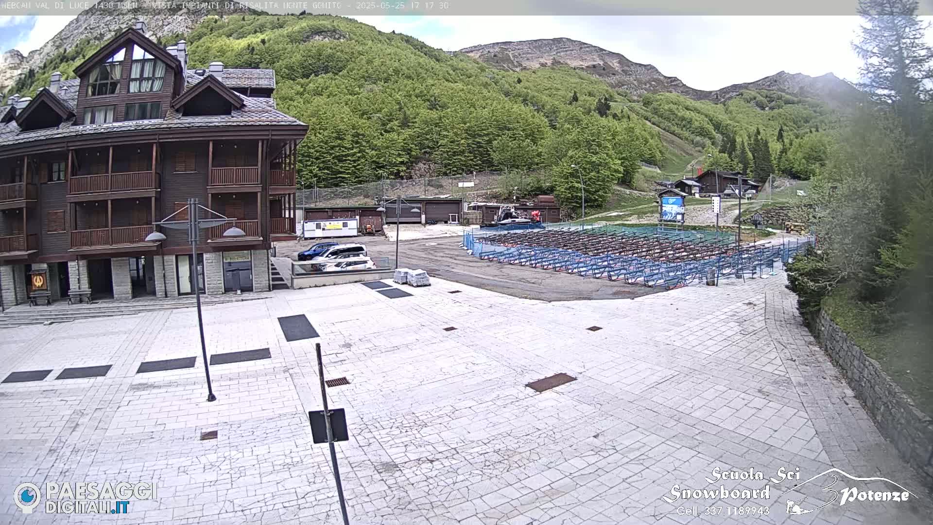 A mostly empty paved plaza sits before a multi-story brown building, with numerous rows of folded metal equipment in the background against a backdrop of a verdant mountainside under a partly cloudy sky.