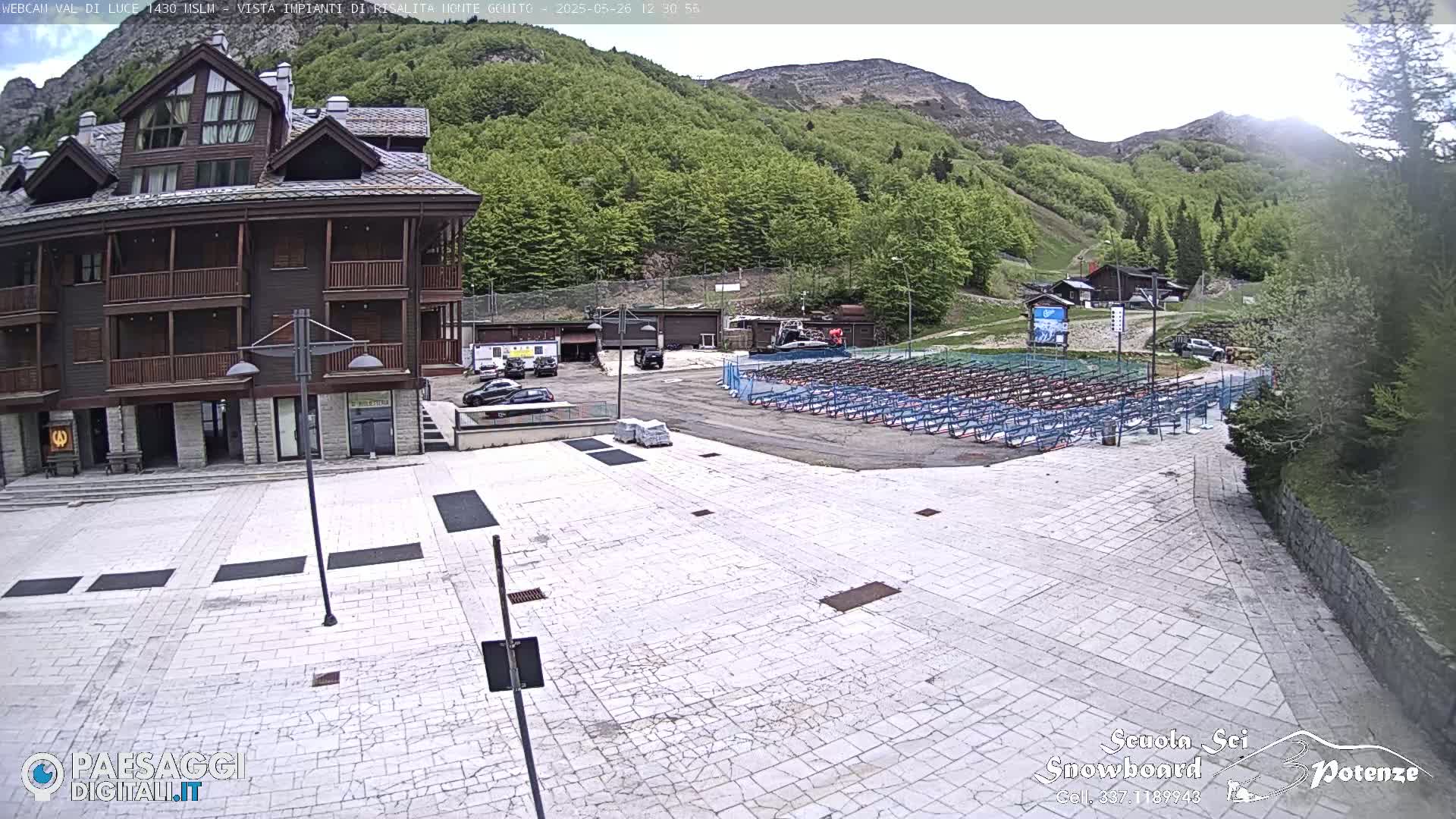 A large paved plaza sits in front of a multi-story building, with rows of what appear to be snowboard or ski racks in the background, all set against a backdrop of a mountainside covered in lush green vegetation under a mostly clear sky.