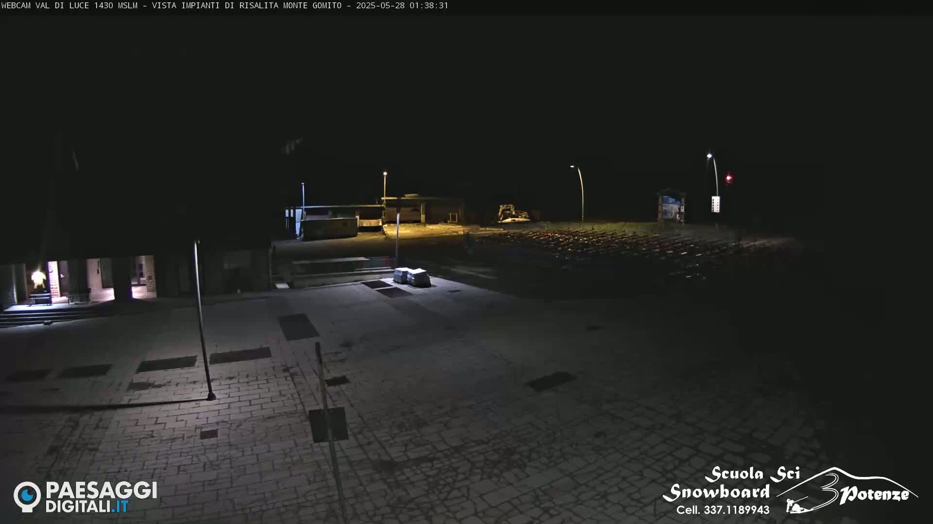 A nighttime, outdoor view of a paved plaza with some buildings and sparsely lit areas, possibly showing light snow or frost on the ground.