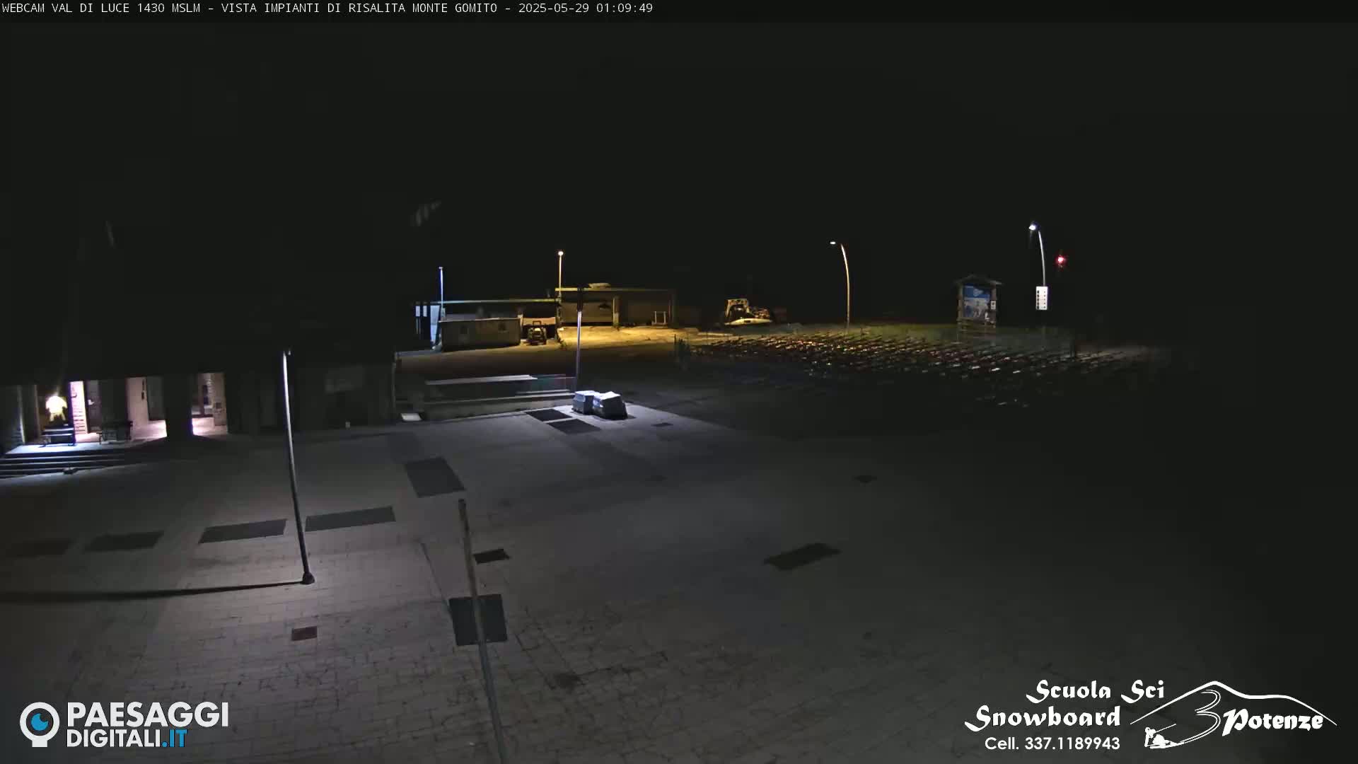 A nighttime, outdoor view of a mostly empty paved plaza with a few buildings and a large parking lot in the background.