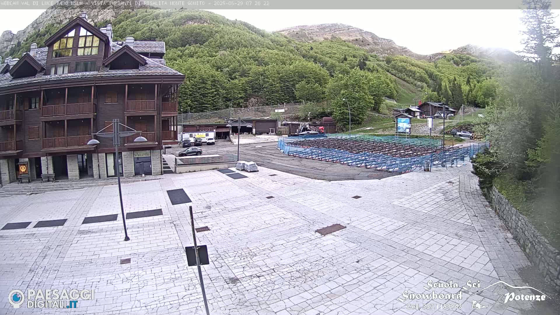 A stone plaza in front of a multi-story brown building is partially filled with numerous rows of stacked snowboards and other equipment, with a mountainside covered in green trees visible in the background under a partly cloudy sky.