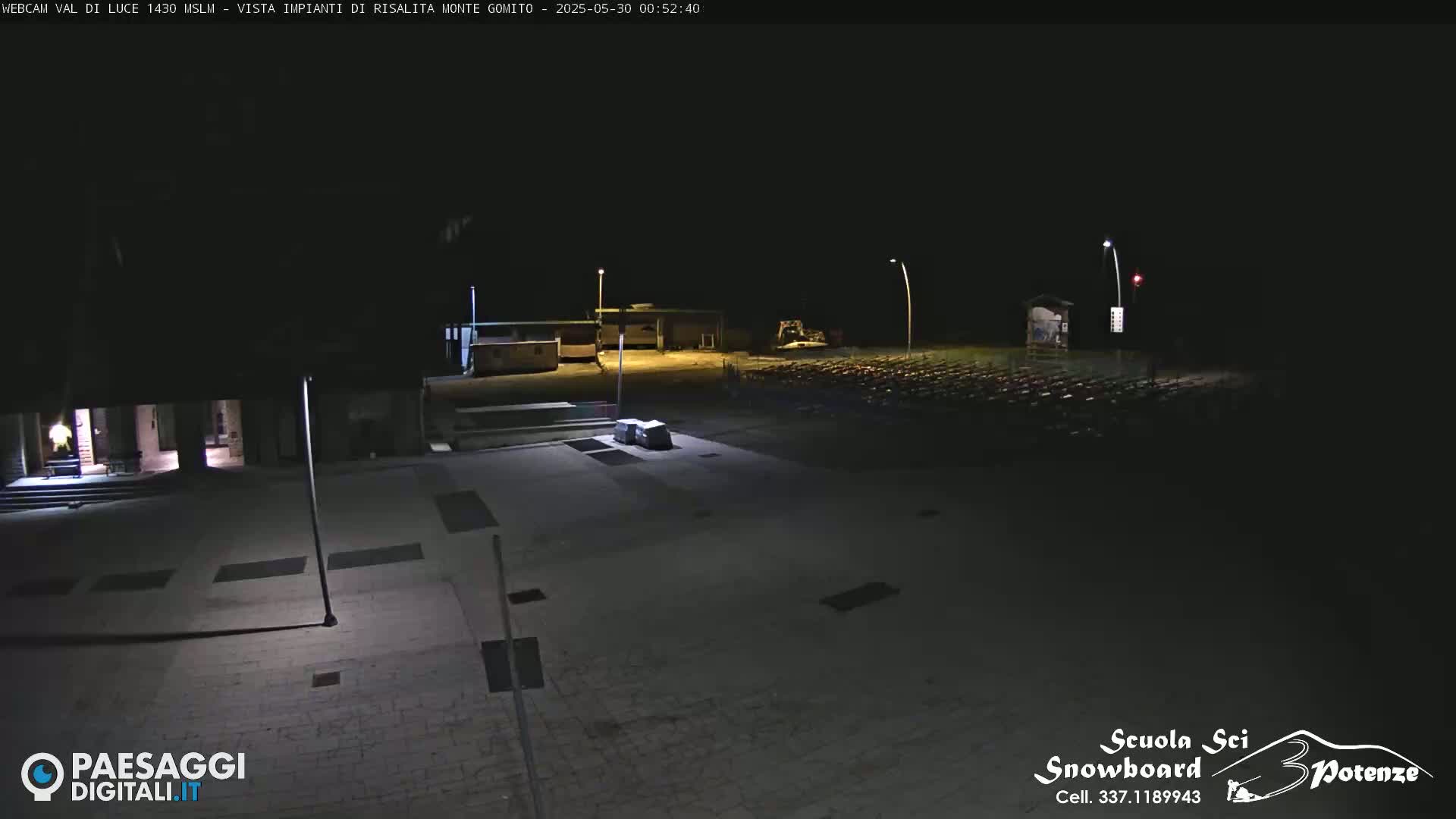 A nighttime view of an empty paved plaza with a few buildings and a large area of what appears to be folded chairs or similar objects in the distance.