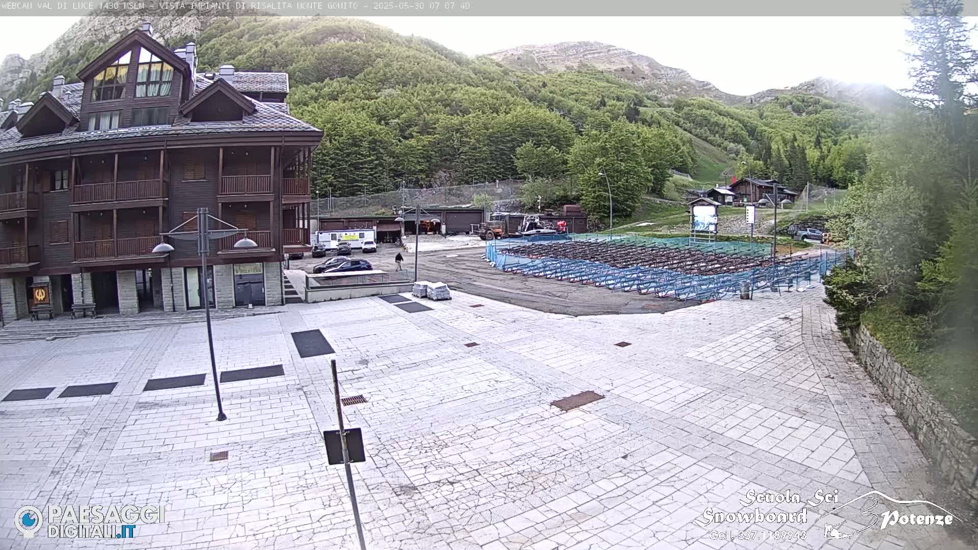 A large, light-colored paved plaza sits in front of a brown multi-story building, with rows of what appears to be ski or snowboard racks in a parking area behind it, all set against a backdrop of a mountainside covered in lush green trees under an overcast sky.