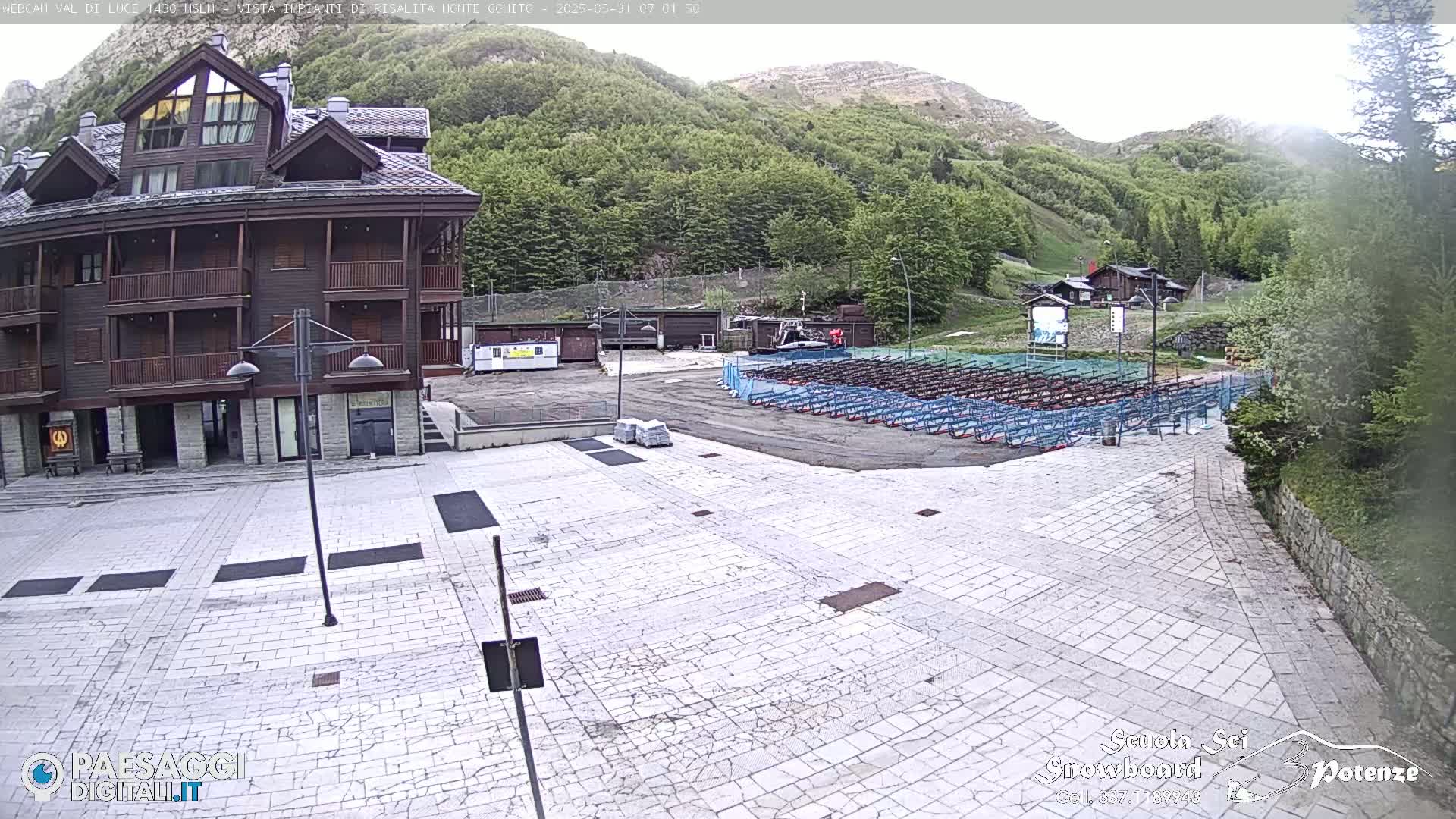 A mostly empty paved plaza sits in front of a multi-story brown building, with rows of what appears to be snow sport equipment storage in the background under an overcast sky.