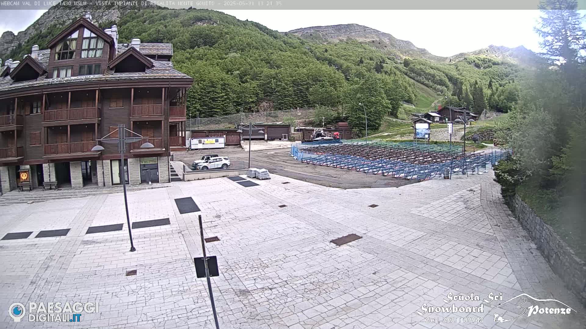 A large, light-colored paved plaza sits in front of a multi-story brown building, with a mountainside covered in green trees visible in the background under a mostly clear sky.