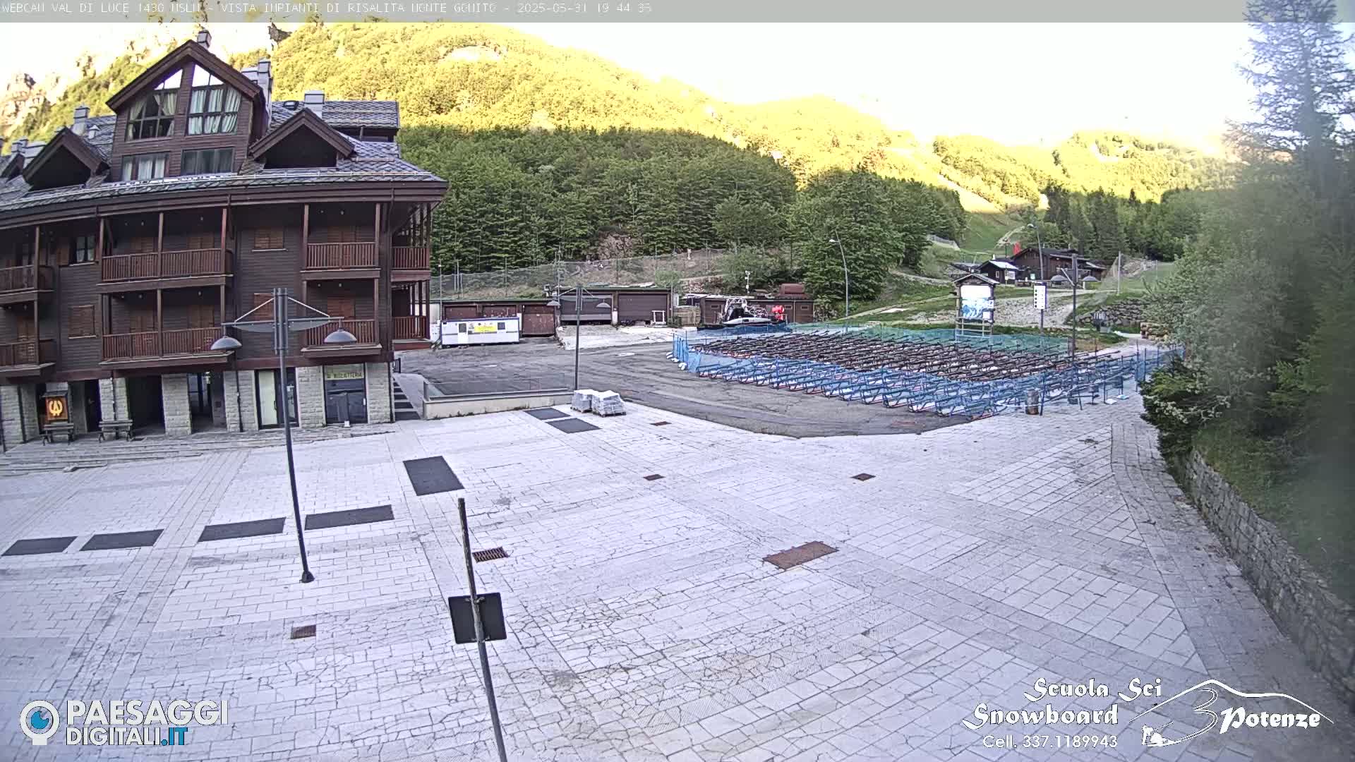A stone plaza sits in front of a multi-story wooden building, with a mountainside covered in green trees in the background under a bright, sunny sky.