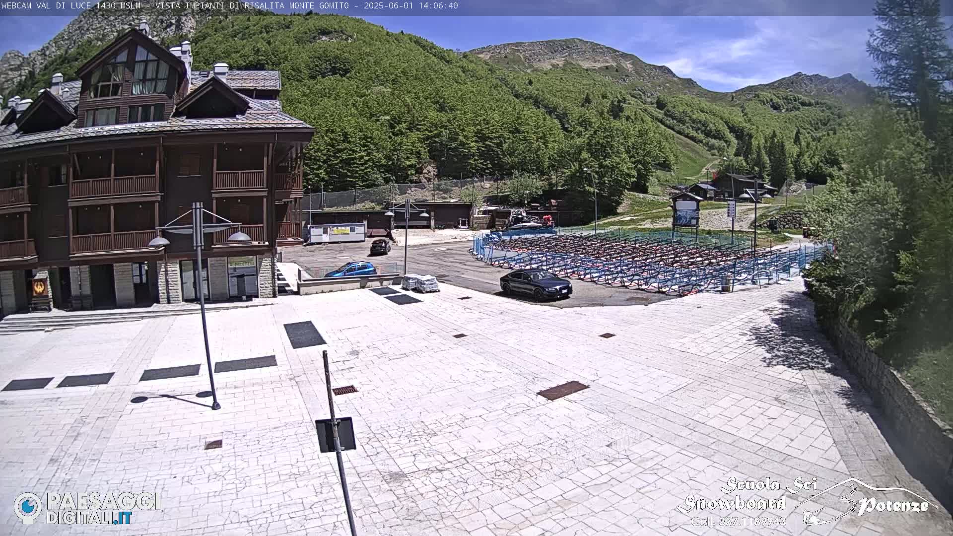 A paved plaza with a few cars is situated in front of a multi-story building, overlooking a mountainside covered in lush green trees under a partly sunny sky.