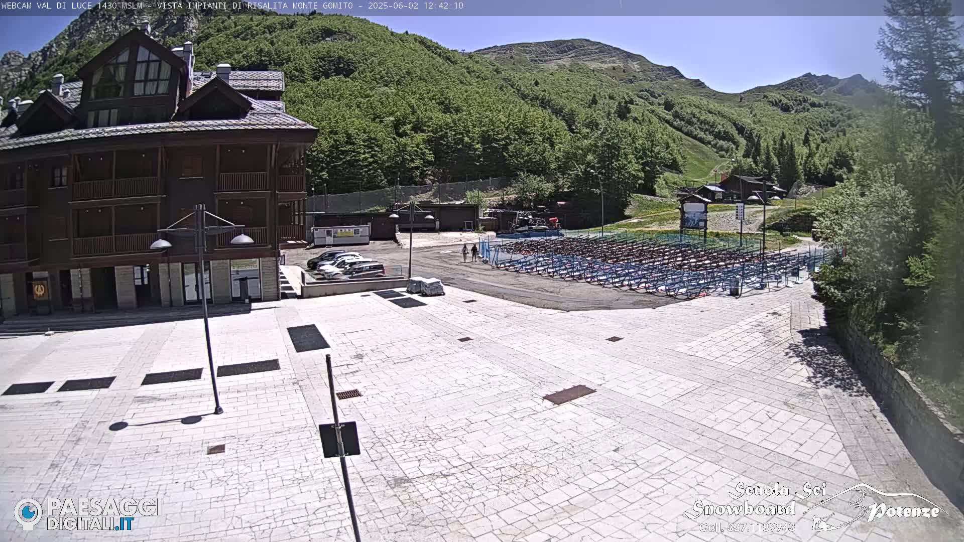 A stone plaza sits before a brown multi-story building, with a large area of folded seating and a mountain backdrop under a sunny sky.