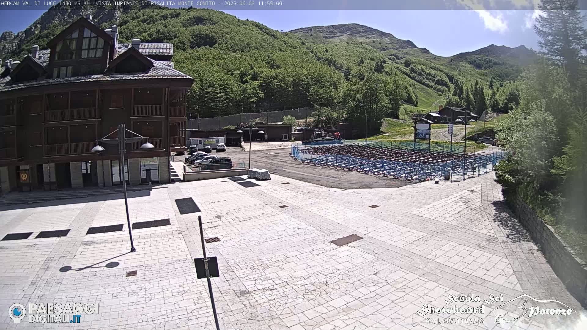 A paved plaza sits before a multi-story brown building, with a large area of folded metal chairs in the background under a sunny sky and green, mountainous terrain.