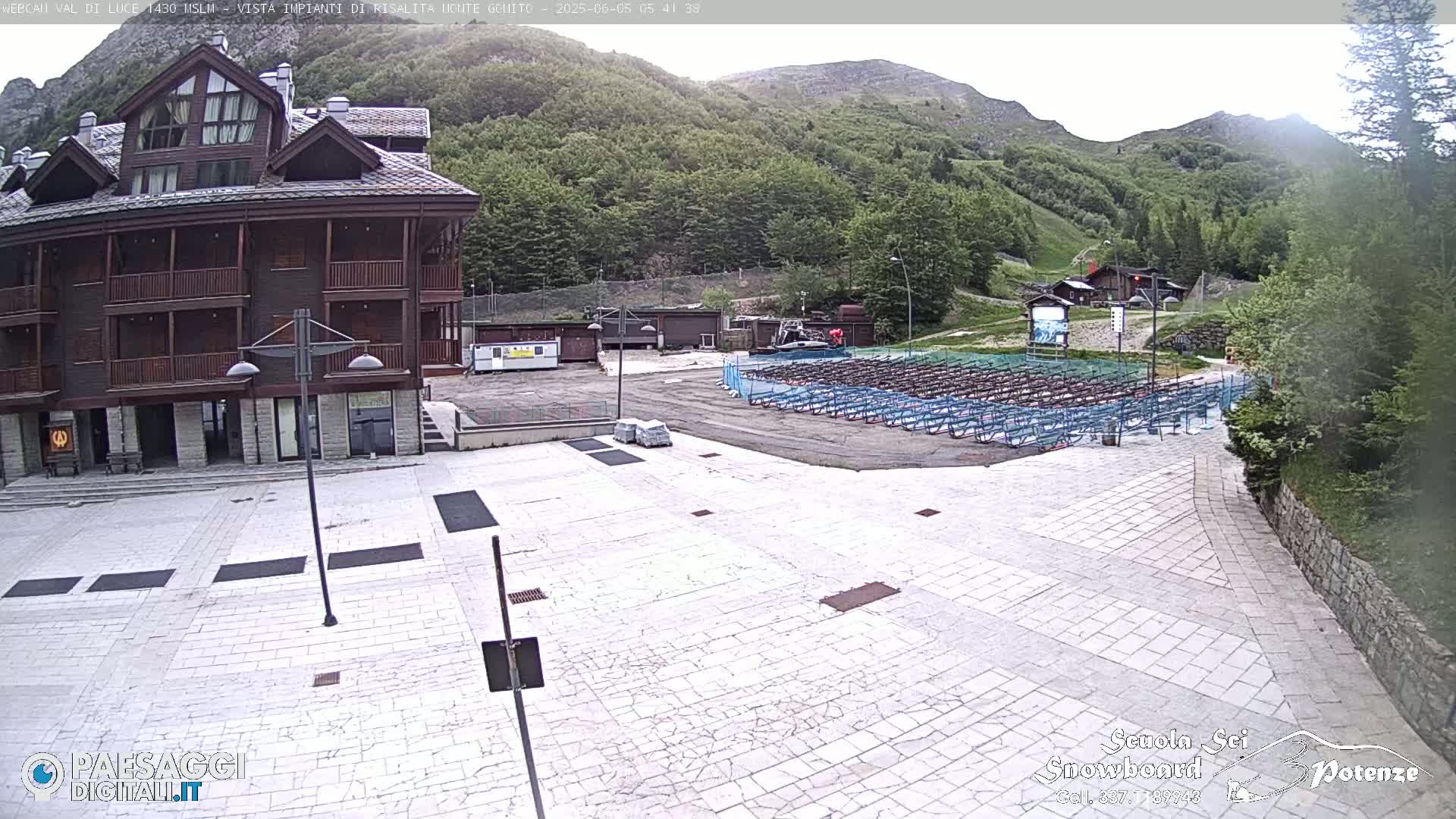 A stone plaza sits in front of a multi-story wooden building, with rows of folded metal seating and a mountainside in the background on an overcast day.