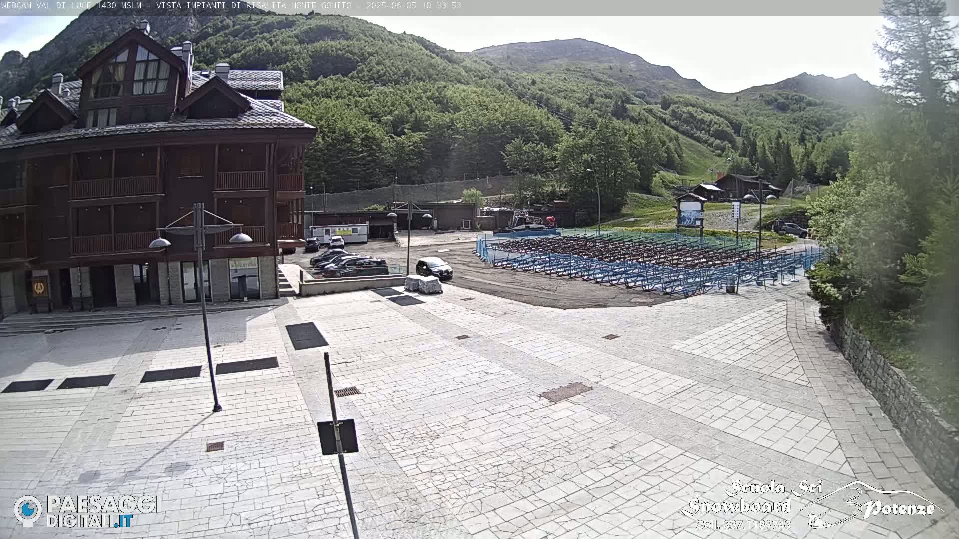 A large paved plaza sits before a multi-story wooden building, with a mountainside covered in lush green trees and a row of folded chairs in the background under a sunny sky.