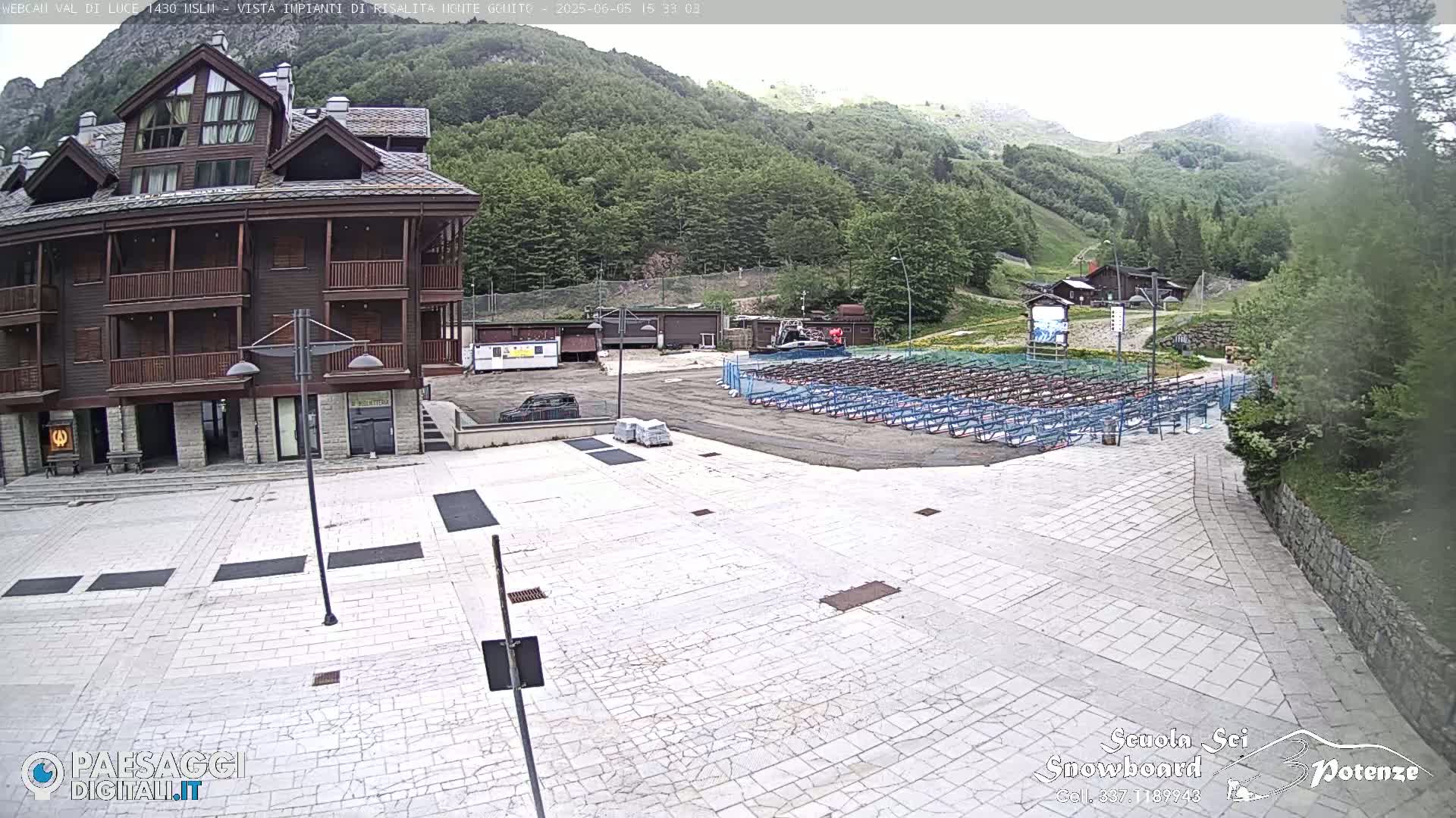 A mostly empty paved plaza sits in front of a multi-story brown building, with a mountainside covered in green trees visible in the background under an overcast sky.