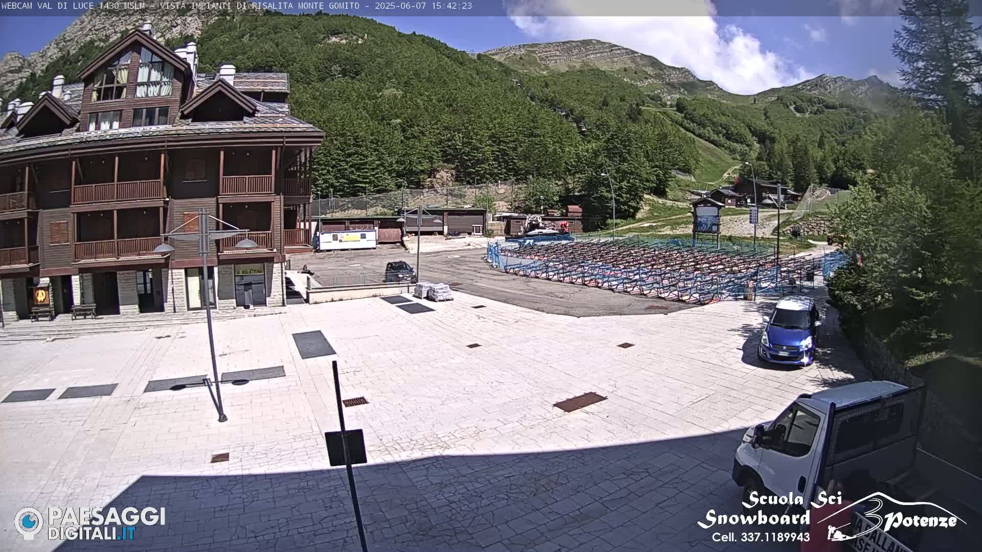 A paved plaza sits before a multi-story wood-sided building, with a mountainside covered in green trees and a partly cloudy sky in the background.
