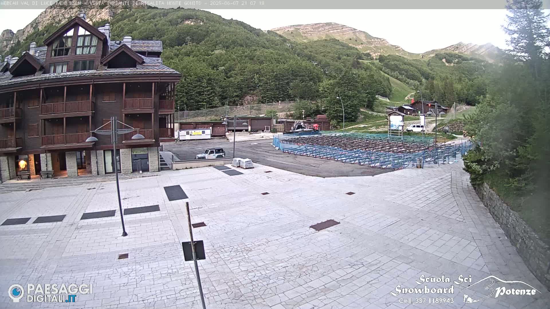 A large, paved plaza sits in front of a three-story brown building, with rows of metal equipment and a mountainside visible in the background on a partly cloudy day.