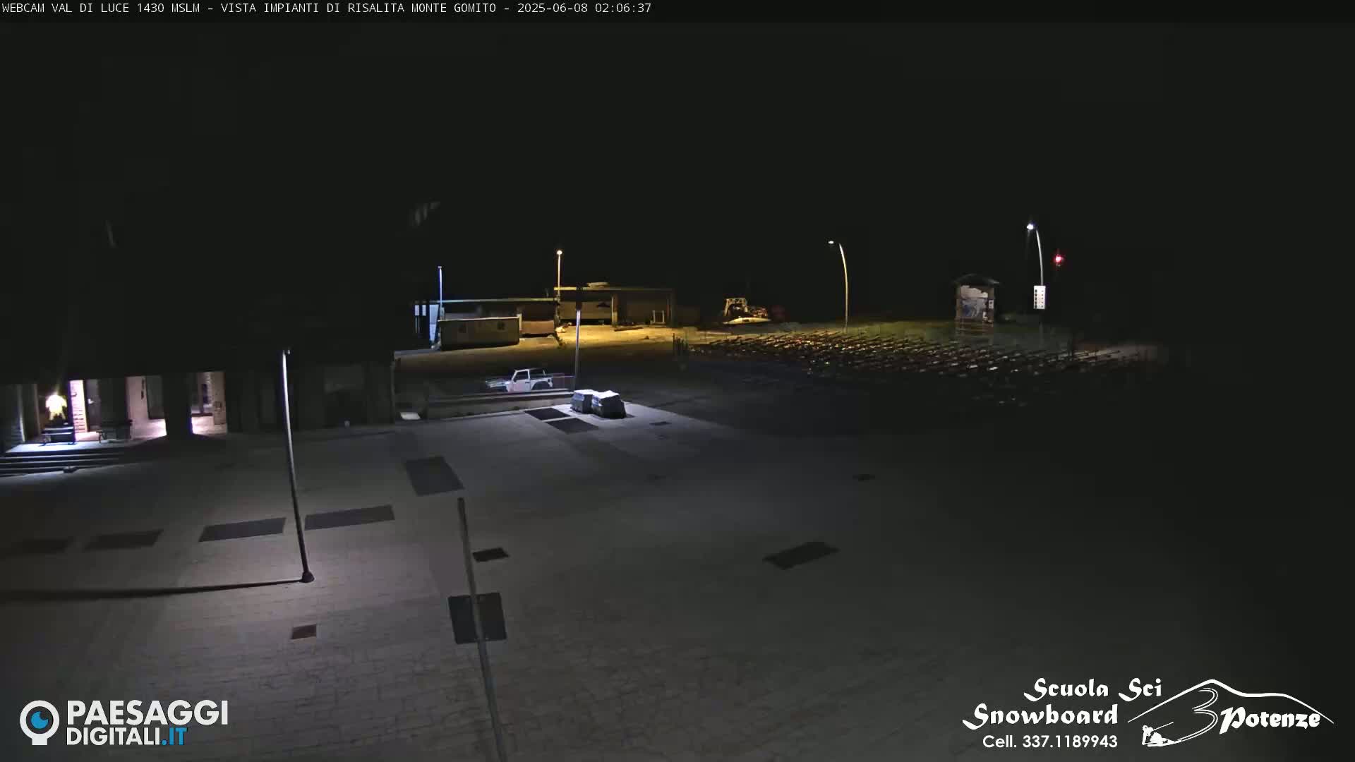 A nighttime, outdoor view of a mostly empty paved area with a few vehicles, some buildings in the background, and rows of what appear to be outdoor chairs or seating.