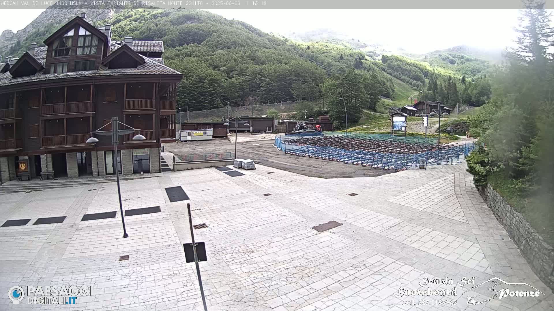 A paved plaza in front of a multi-story wood-sided building, with an empty seating area and green hills in the background on an overcast day.