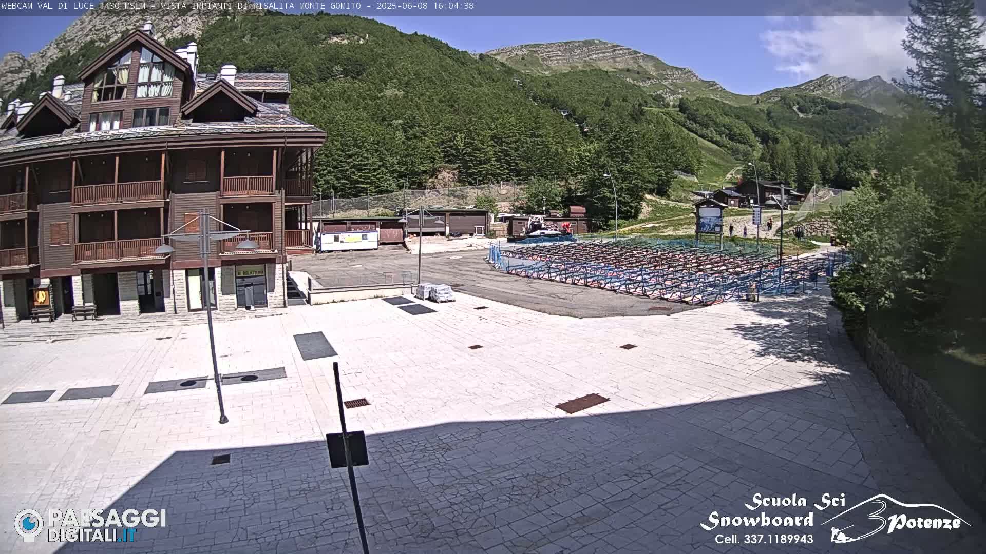 A sunny view of a plaza with a multi-story wooden building, a large area of folded metal structures, and a mountainside in the background.