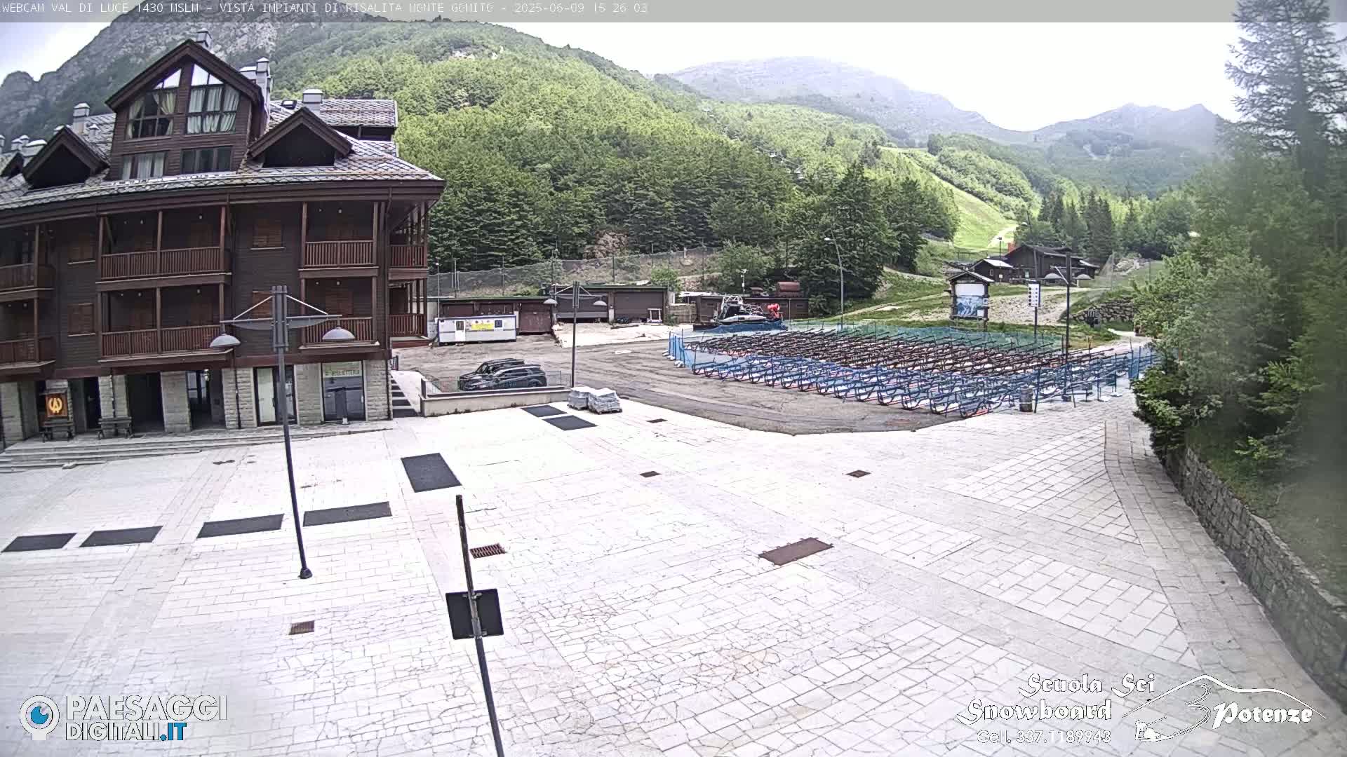 A large paved plaza sits in front of a brown multi-story building, with a mountain backdrop and overcast skies; numerous metal racks are visible in the distance.