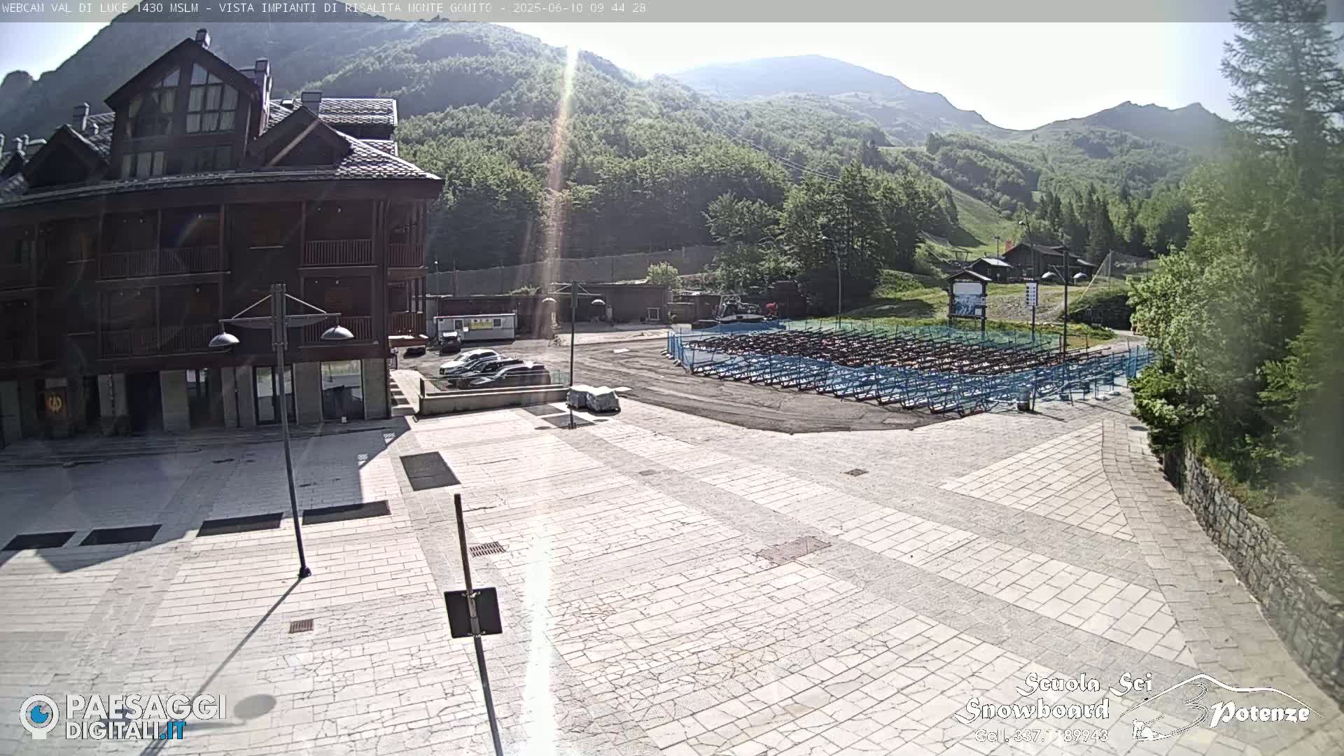 A sunny day reveals a paved plaza in front of a multi-story wooden building, overlooking a hillside with numerous rows of folded seating.