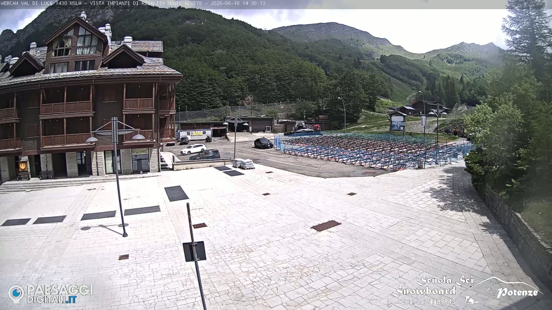 A light-grey paved plaza sits before a multi-story brown building, with a parking area and rows of folded chairs beyond it, all set against a backdrop of green mountains under a partly cloudy sky.