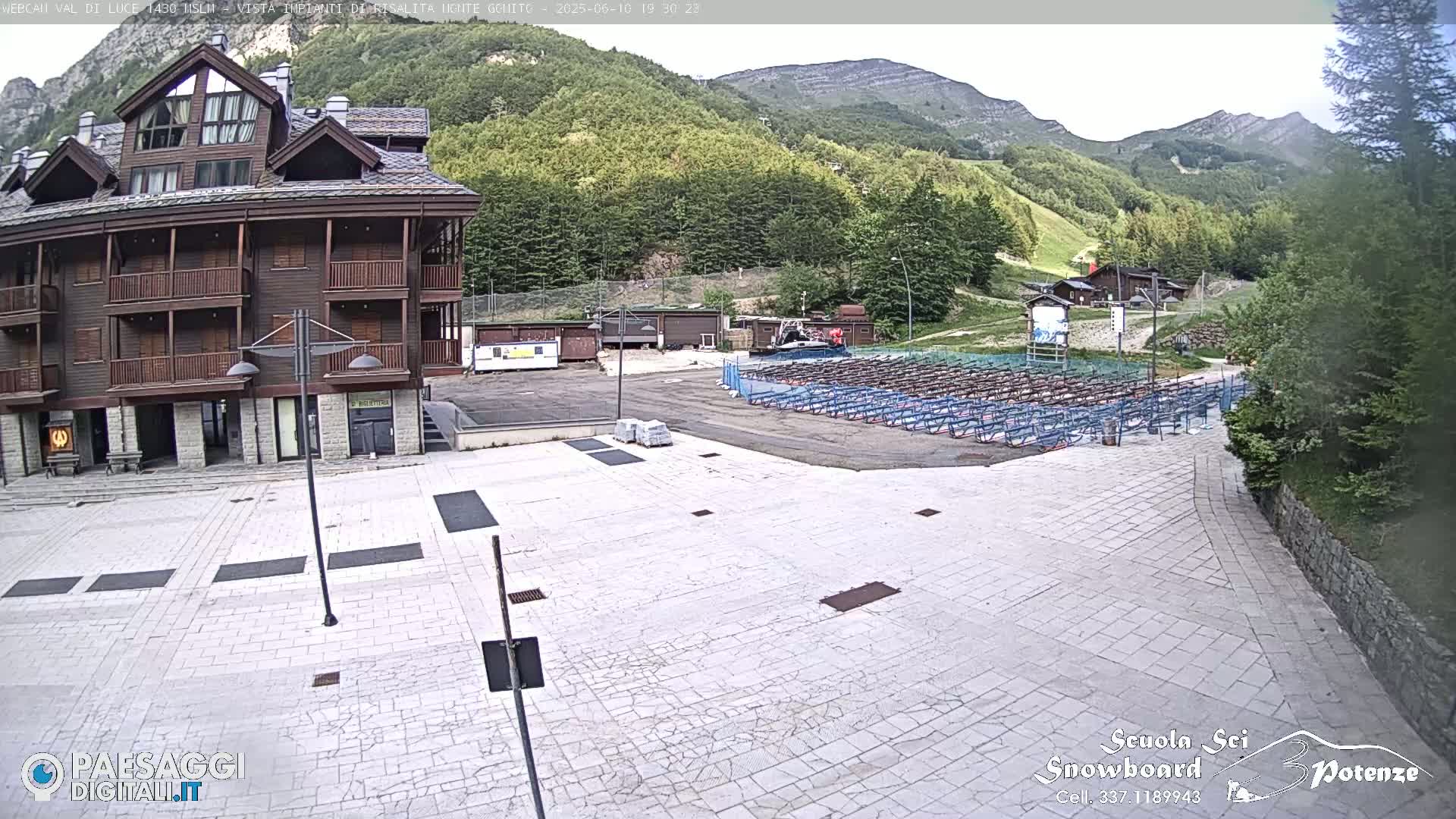 A large, light-colored paved plaza sits in front of a multi-story brown building, with mountains and a ski resort in the background under a partly cloudy sky.
