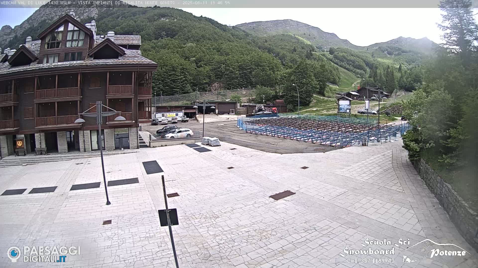 A paved plaza sits before a multi-story wood-and-stone building, with a mountainside and rows of folded seating in the background under a partly cloudy sky.