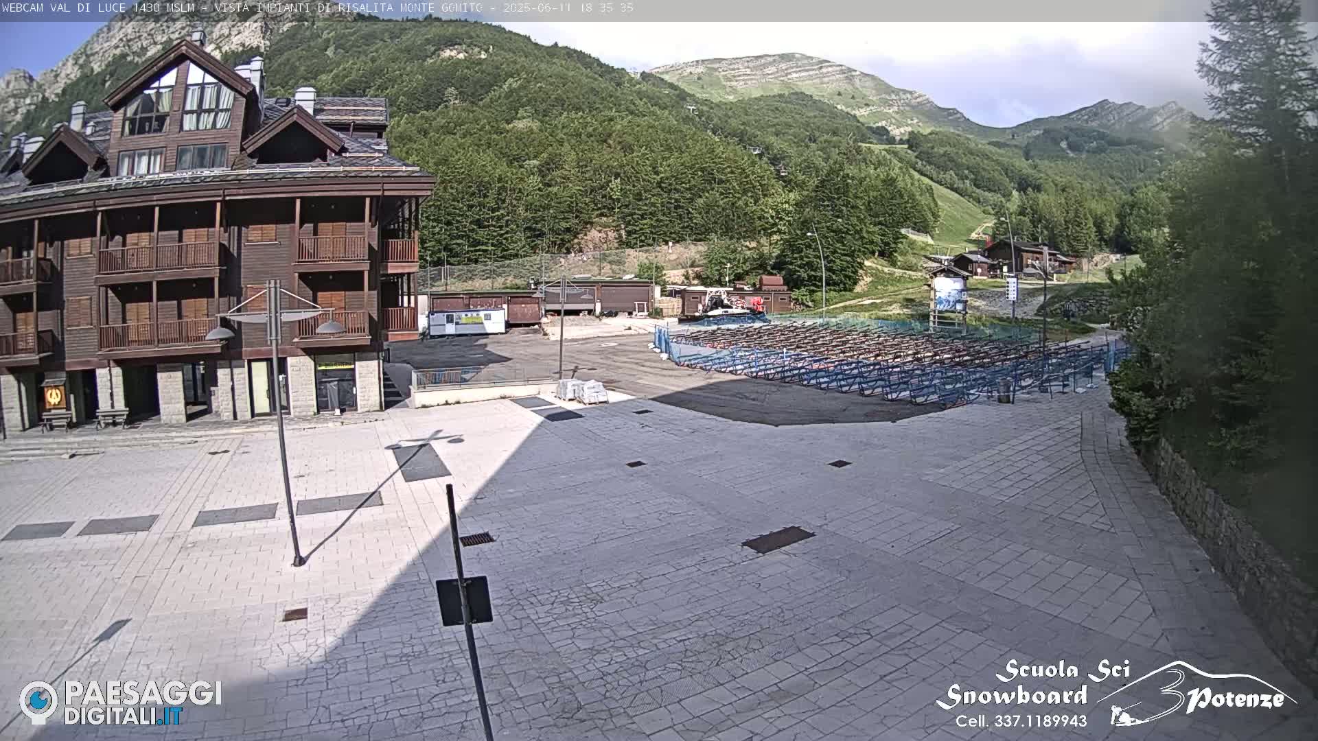 A mostly sunny view of a paved plaza, a multi-story wood building, and a mountainside with ski lift infrastructure in the background.