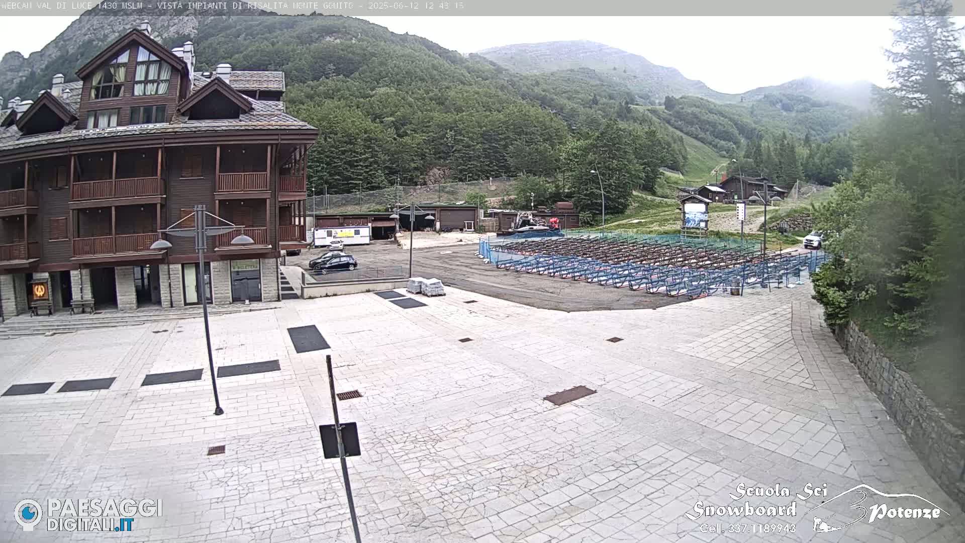 A large, light-colored paved plaza sits in front of a multi-story wood-sided building, with mountains and a forest visible in the background under a mostly cloudy sky.