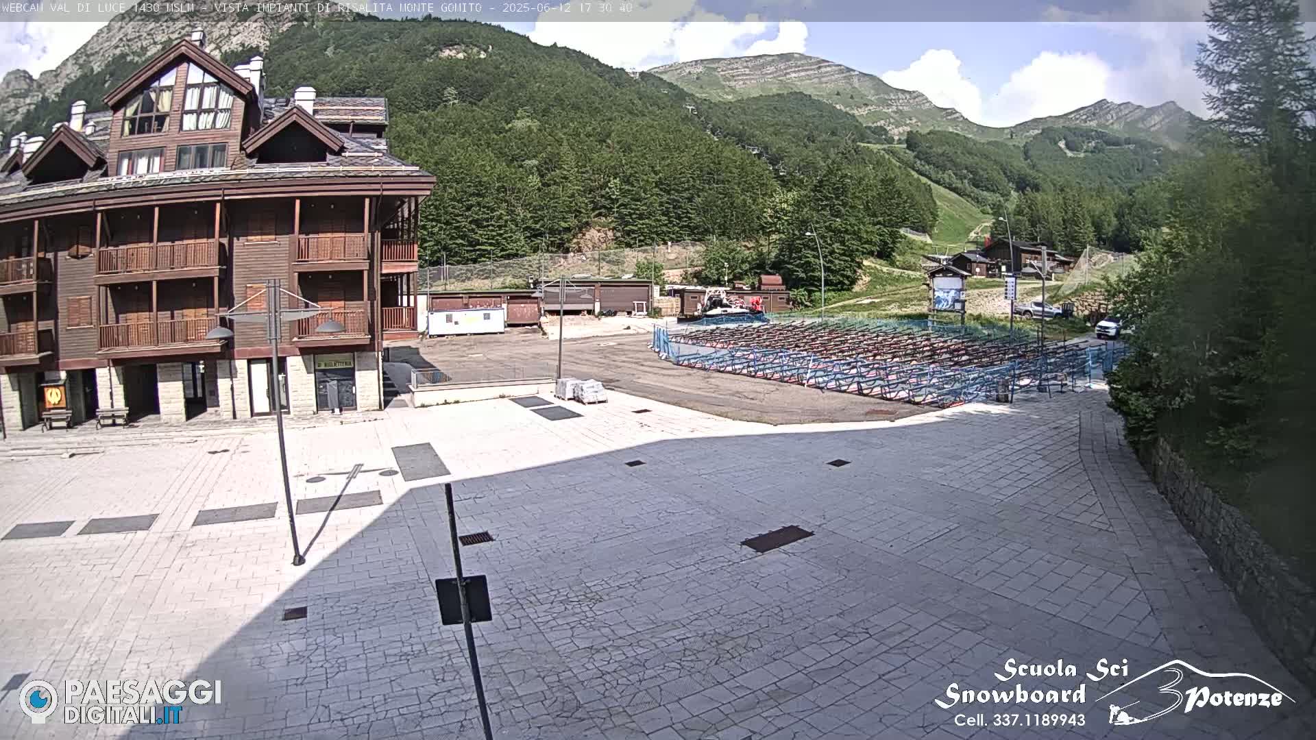 A paved plaza sits before a multi-story wooden building, with a mountain range visible in the background under partly cloudy skies.