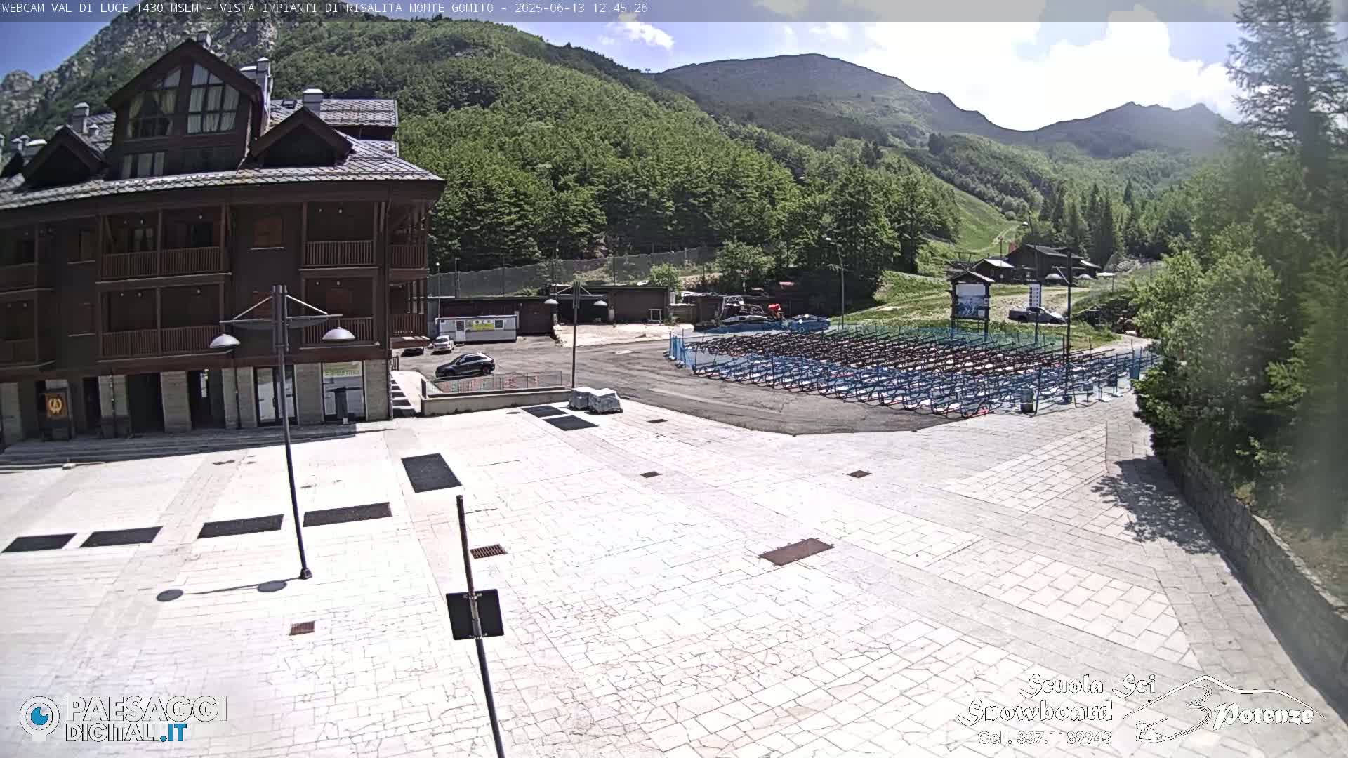 A paved plaza is in front of a multi-story building, with a large area of folded seating in the background, all set against a backdrop of a mountain range under a partly cloudy sky.