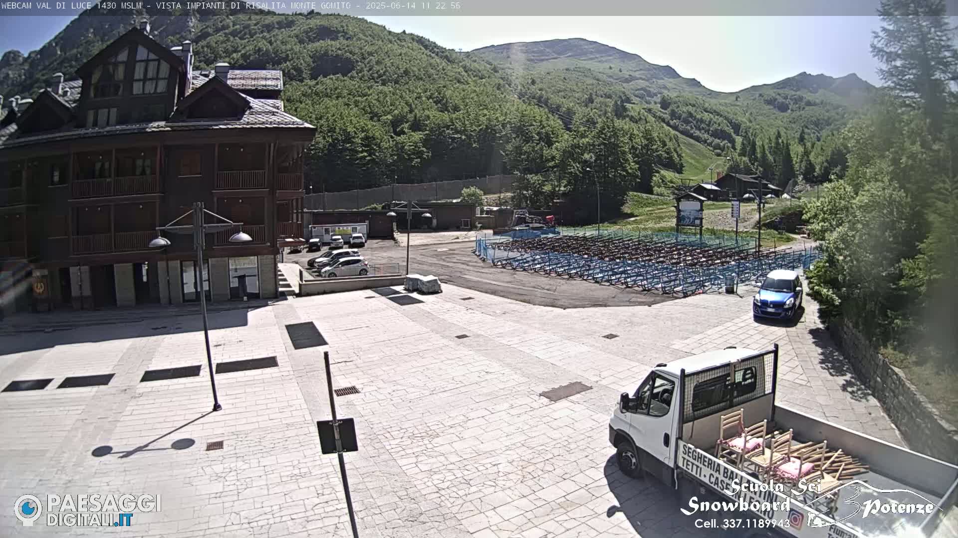 A large, light-colored paved area in front of a multi-story wooden building, with mountains in the background under a sunny sky; a van is parked in the foreground carrying what appear to be chairs.