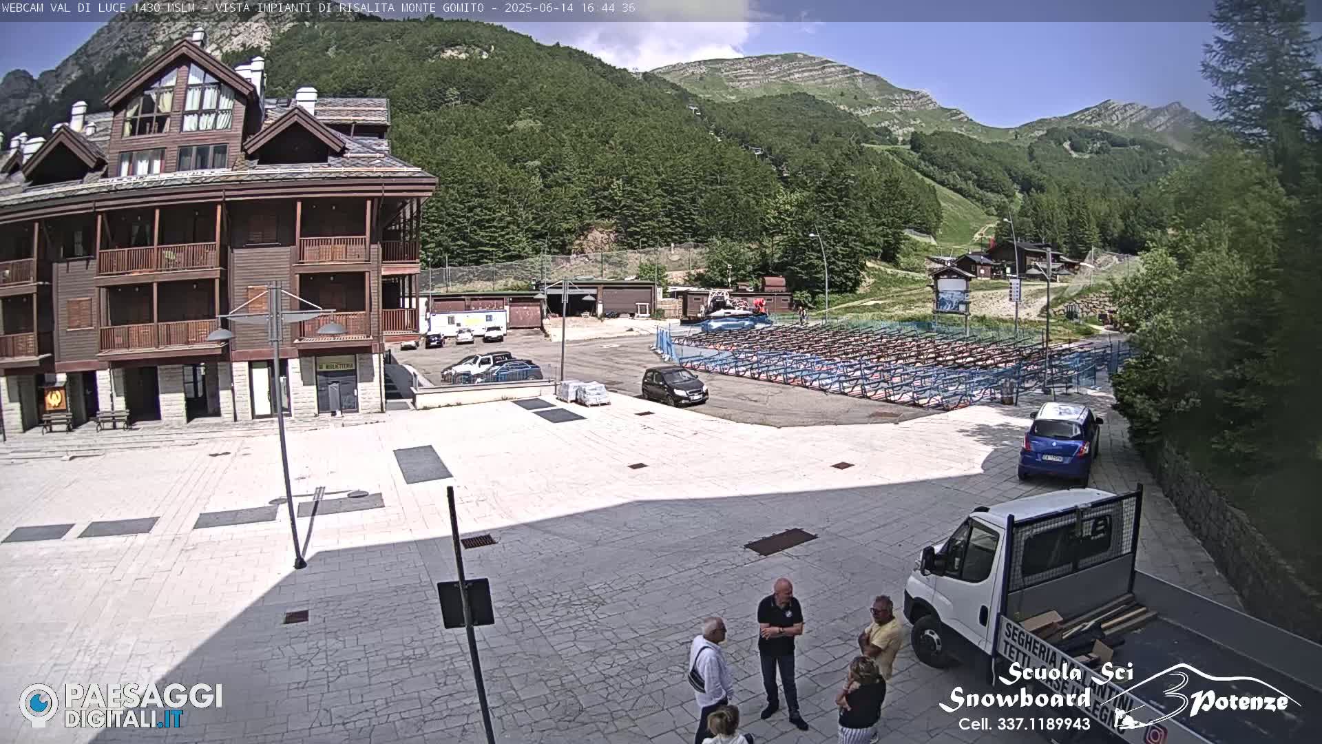 A sunny day reveals a plaza with a multi-story building, parked cars, a group of people conversing, and a mountainside with ski lifts in the background.