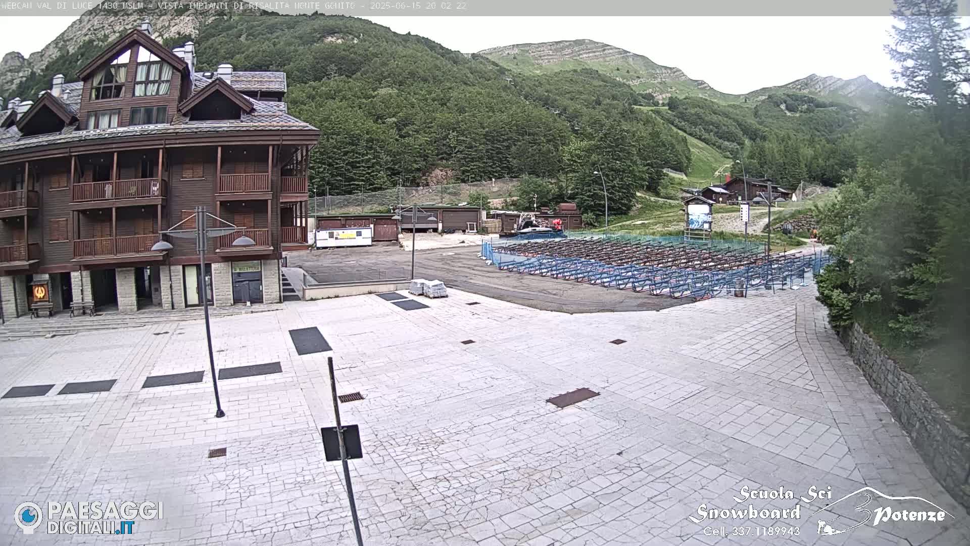 A stone plaza sits in front of a brown multi-story building, with a mountain and forested hills in the background under a partly cloudy sky.