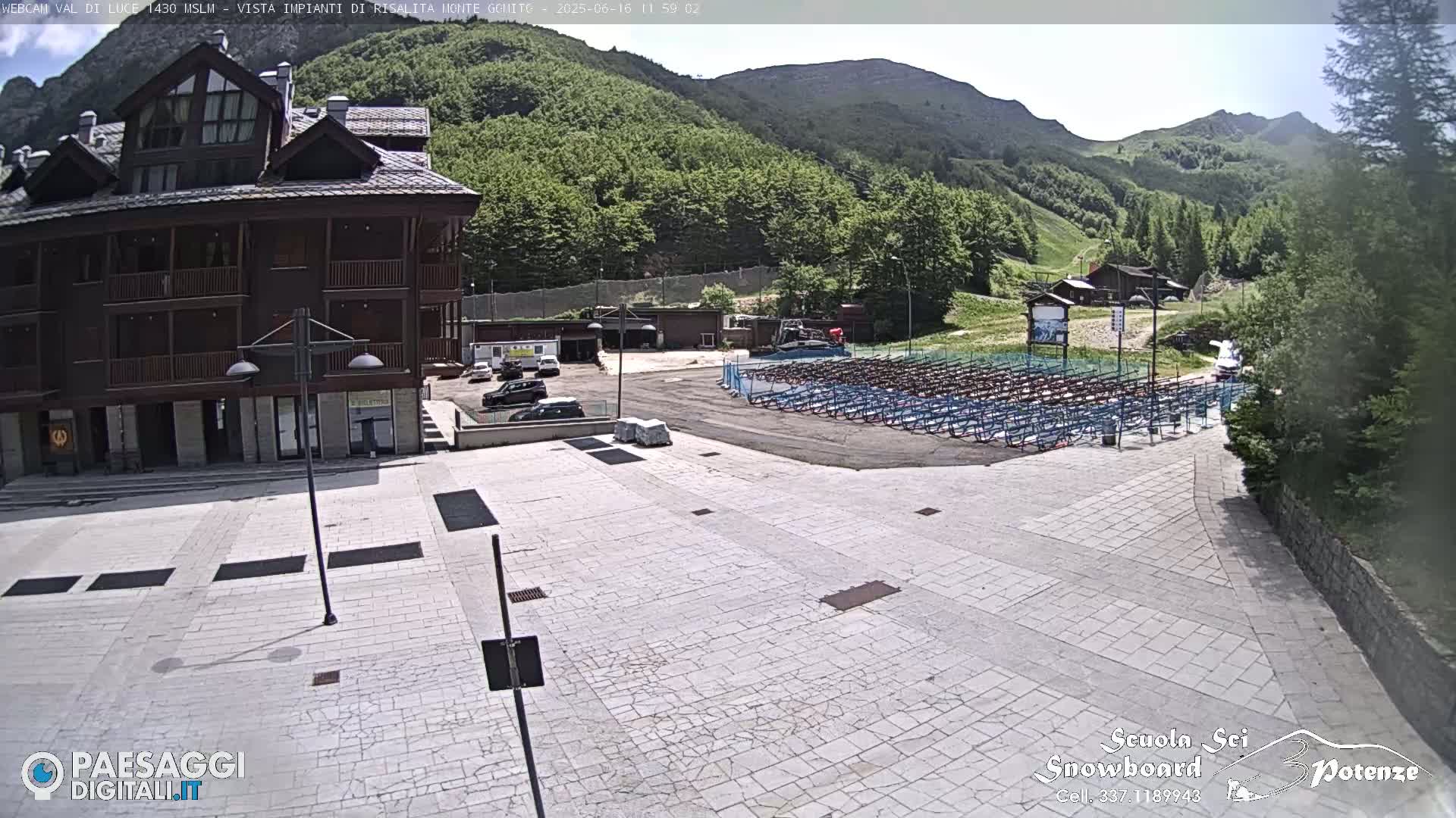 A paved plaza sits in front of a multi-story brown building, with numerous rows of empty chairs in a parking lot beyond it, all under a sunny sky and surrounded by green mountains.