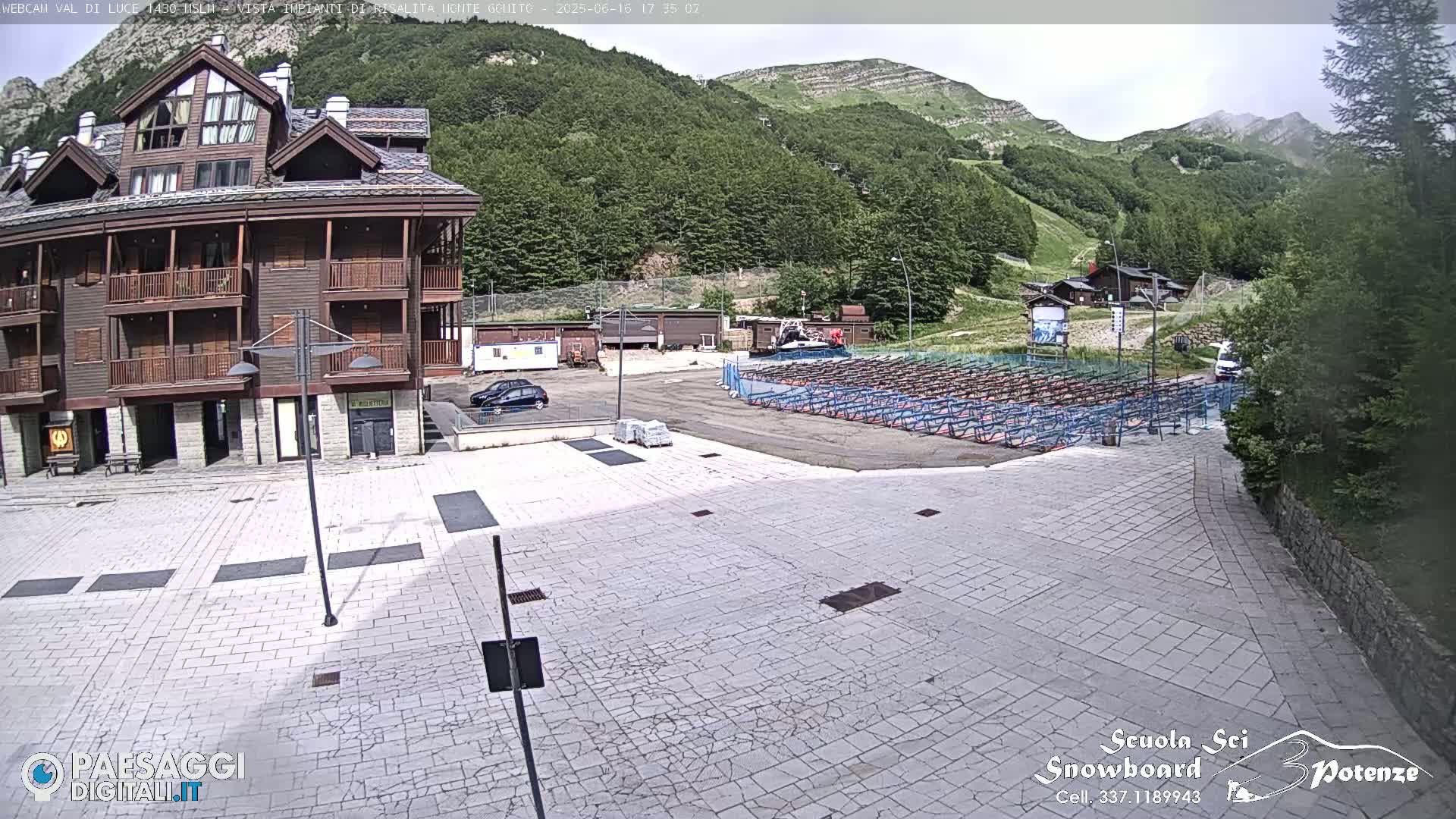 A paved plaza sits before a multi-story wood-sided building, with a mountainside and rows of what appear to be ski racks visible in the background under a partly sunny sky.