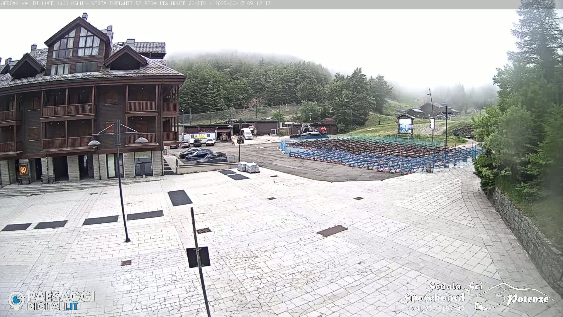 A mostly cloudy day reveals a stone plaza in front of a multi-story wood-sided building, with rows of equipment racks in the midground and a hazy mountainside in the background.