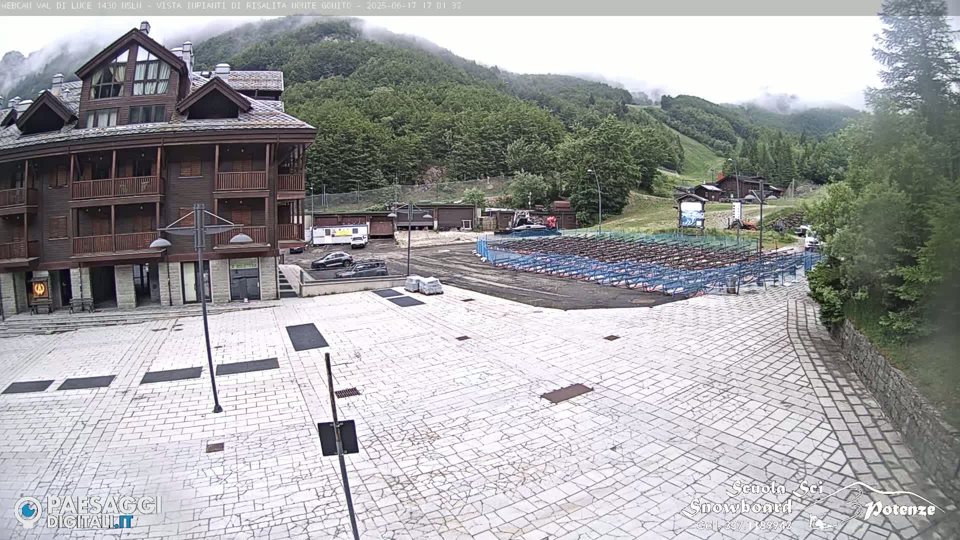 A stone plaza sits in front of a multi-story wood-sided building, with a misty mountain backdrop and numerous rows of folded metal seating in the background.