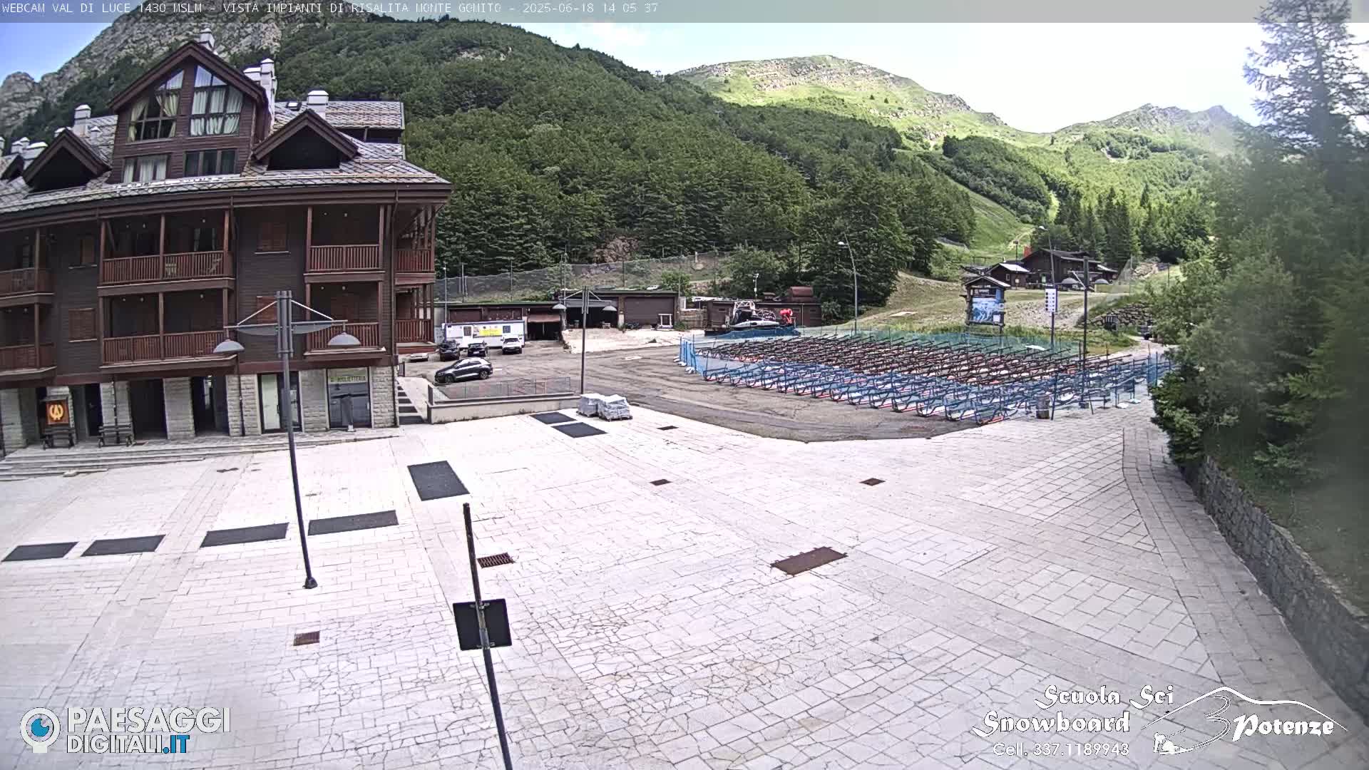 A paved plaza sits before a multi-story wooden building, with a mountainside covered in lush green trees visible in the background under a clear sky.