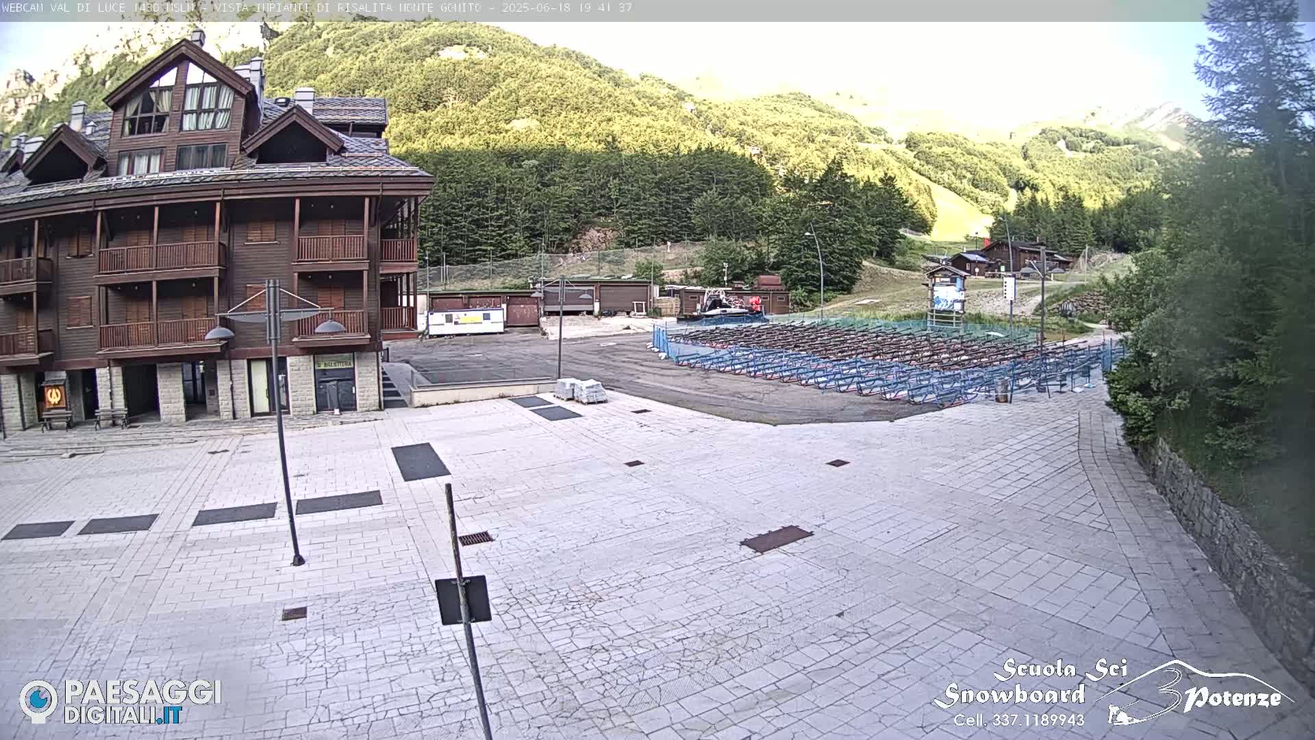 A large paved plaza sits in front of a multi-story wooden building, with a hillside covered in green trees and a seating area in the background under a bright, sunny sky.