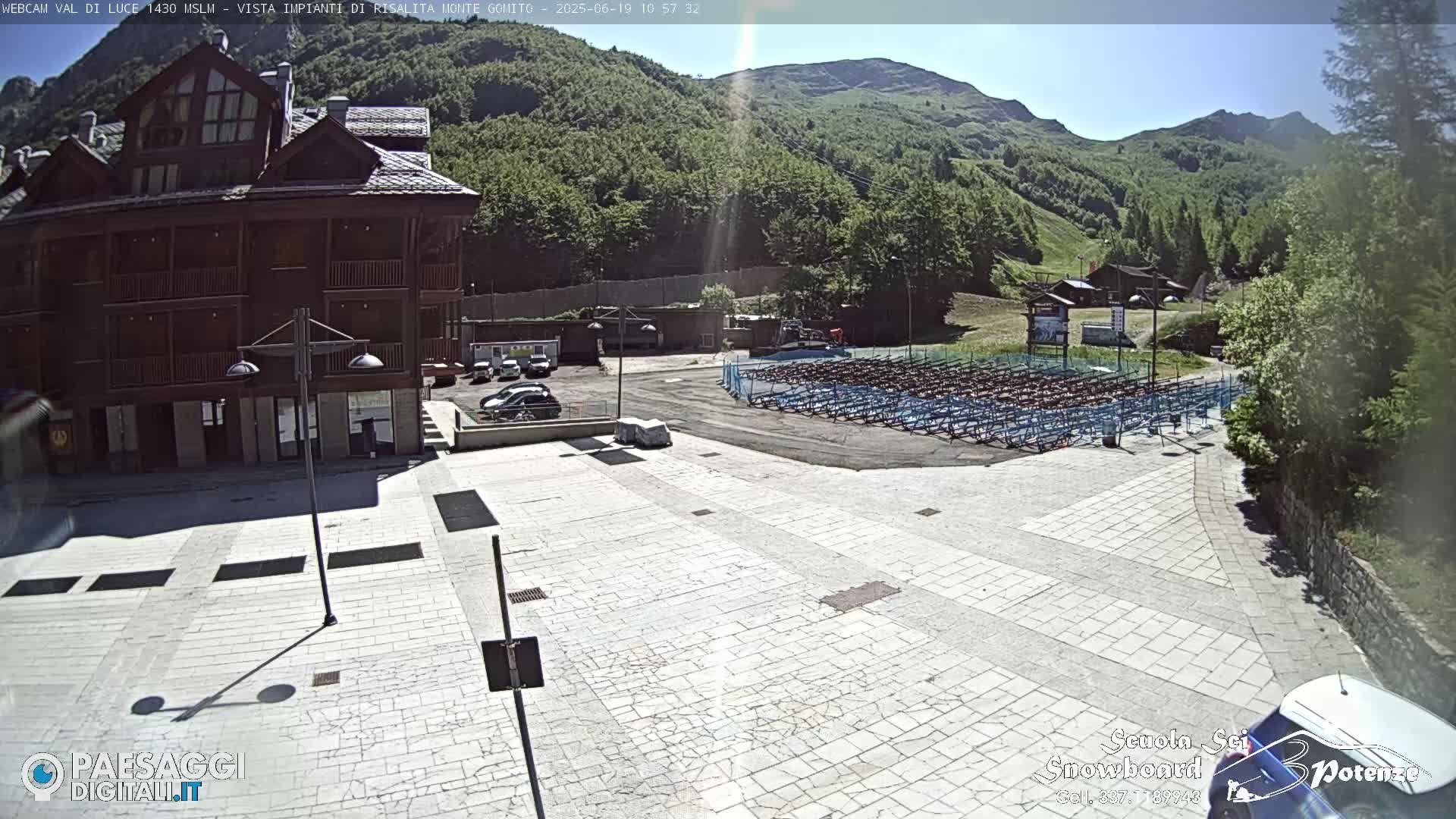 A large paved plaza is situated in front of a multi-story wooden building, with numerous folded chairs in a lot across the plaza, all under a sunny sky against a backdrop of green mountains.