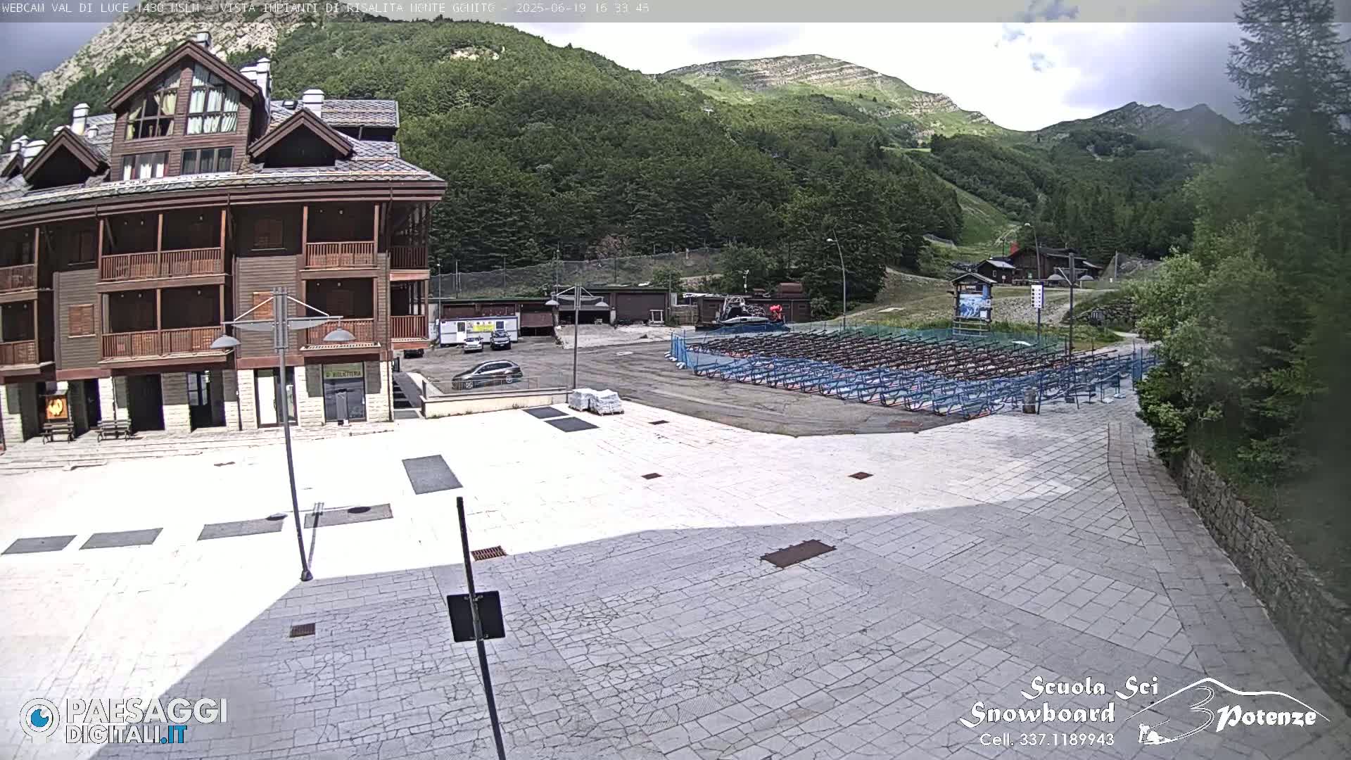 A paved plaza sits before a multi-story wooden building, with a mountain backdrop under a partly cloudy sky.