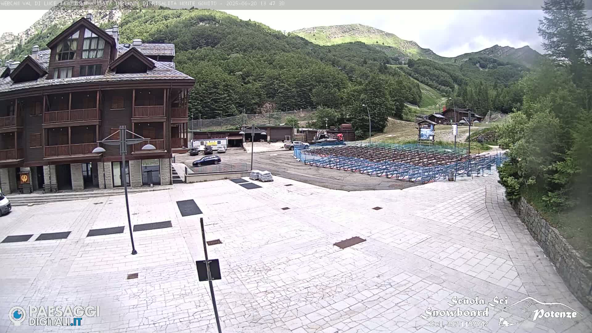 A large paved plaza sits in front of a multi-story brown building, with a mountainside and rows of what appear to be snow sports equipment storage racks in the background, on an overcast day.