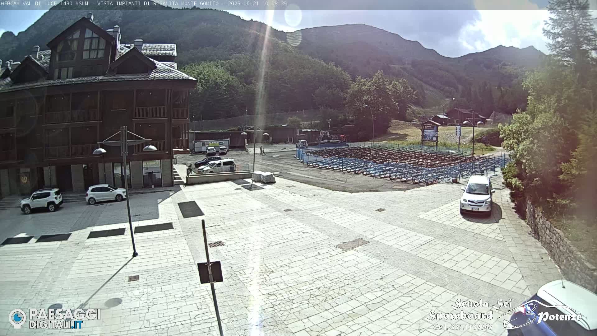 A sunny mountainside plaza with a multi-story building, several parked cars, and a large area of folded seating.