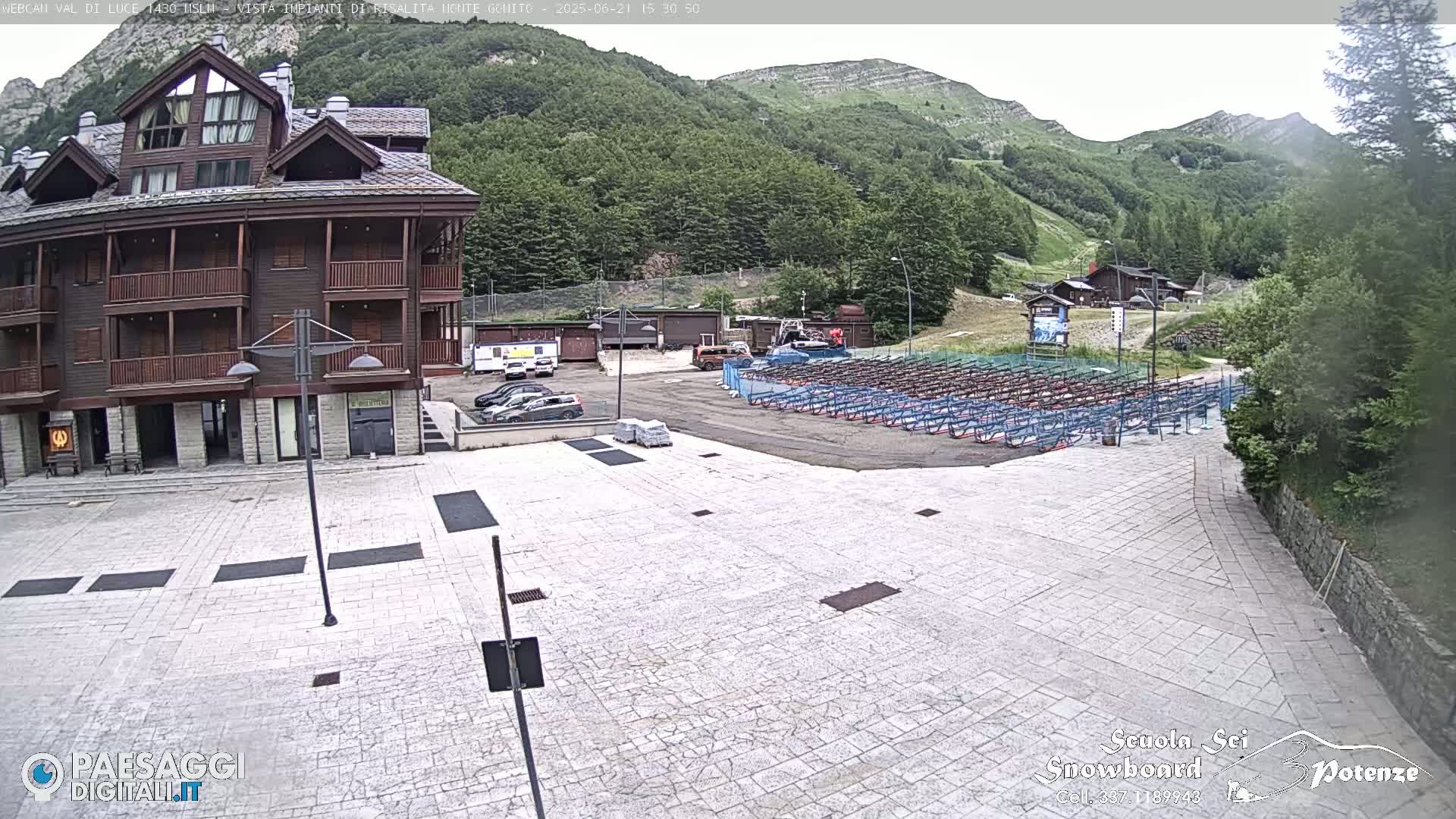 A paved plaza sits before a multi-story brown building, with a mountain backdrop and a large area of stacked equipment visible in the background under a mostly clear sky.
