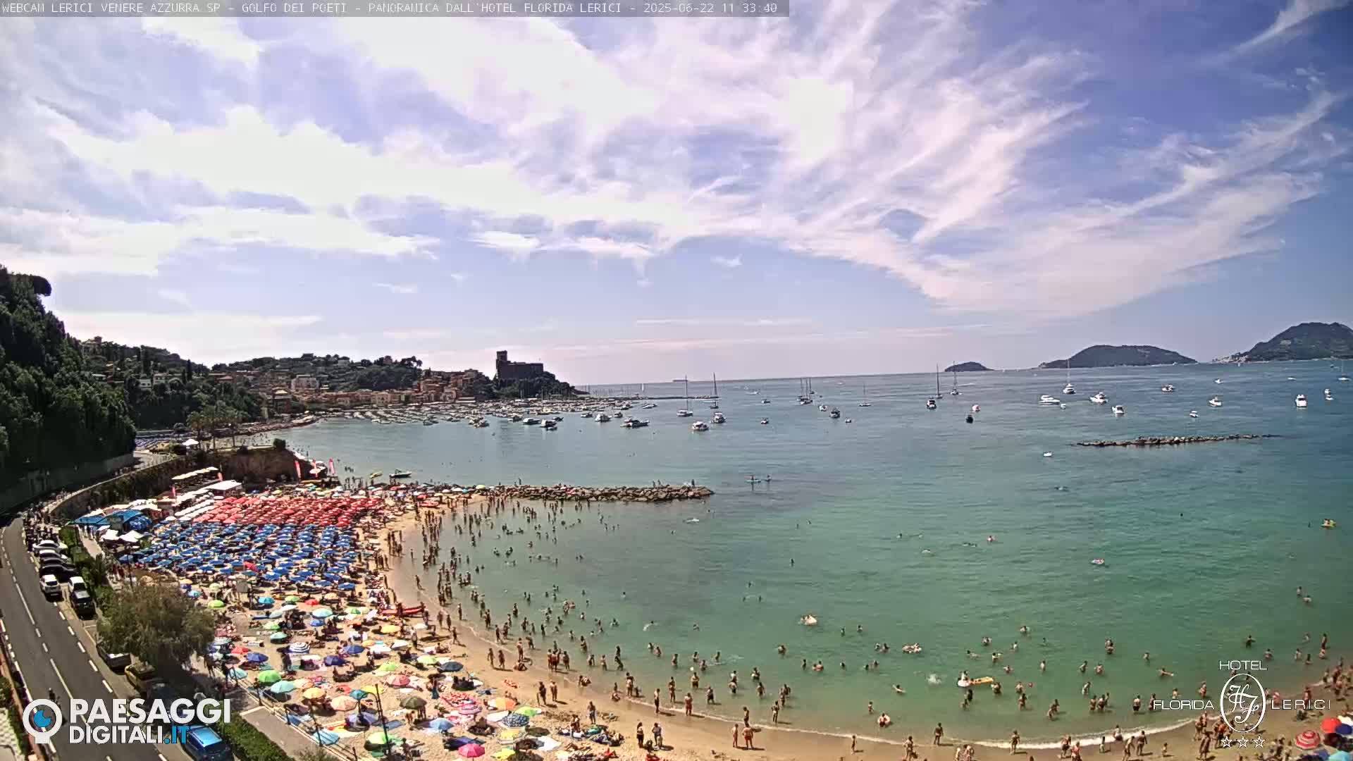 A crowded beach with colorful umbrellas under a partly cloudy sky, boats in a calm bay, and hills in the background.