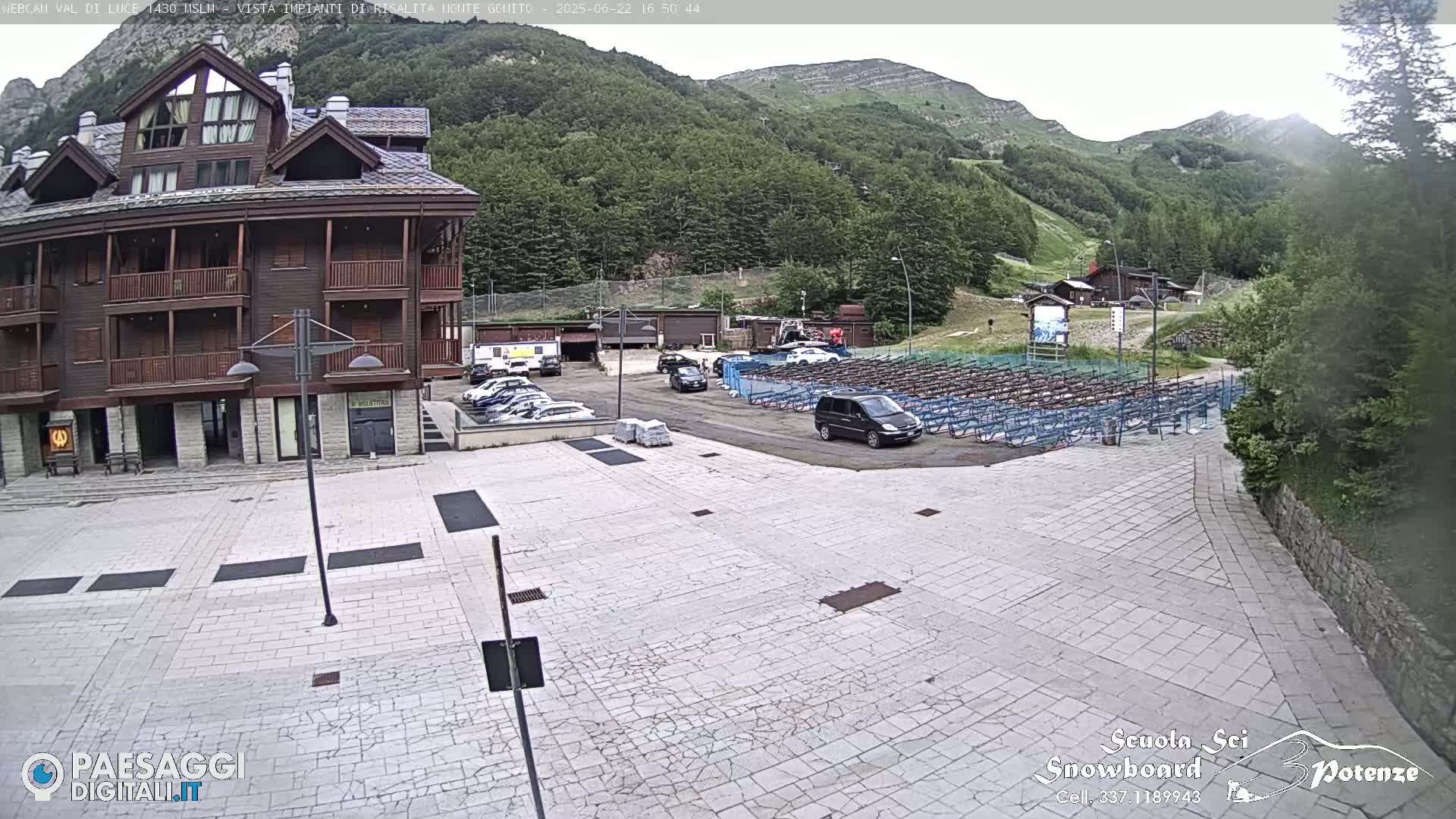 A stone plaza with parked cars in front of a multi-story brown building, overlooked by a mountainside under a partly cloudy sky.