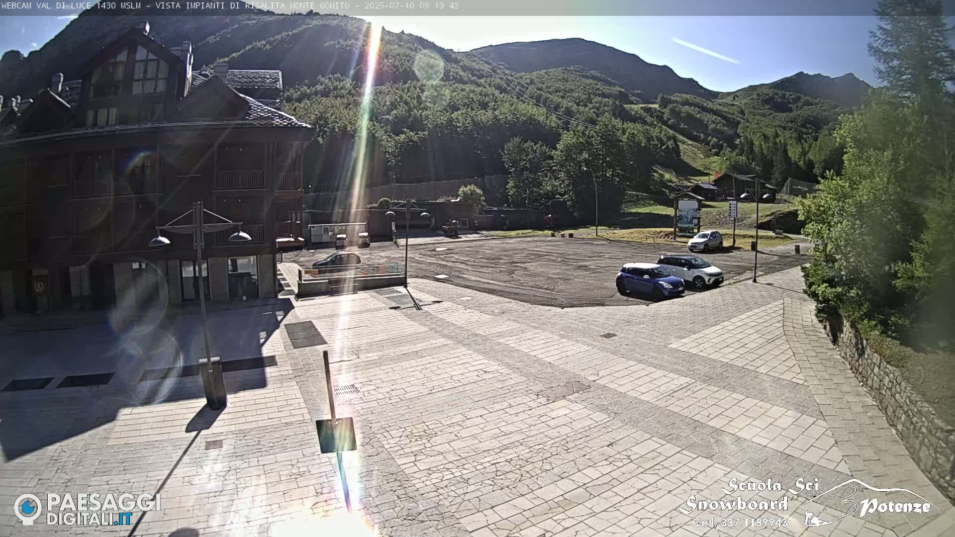 A sunny day reveals a paved plaza with several parked cars in front of a multi-story building, nestled against a backdrop of mountains and lush green hills.
