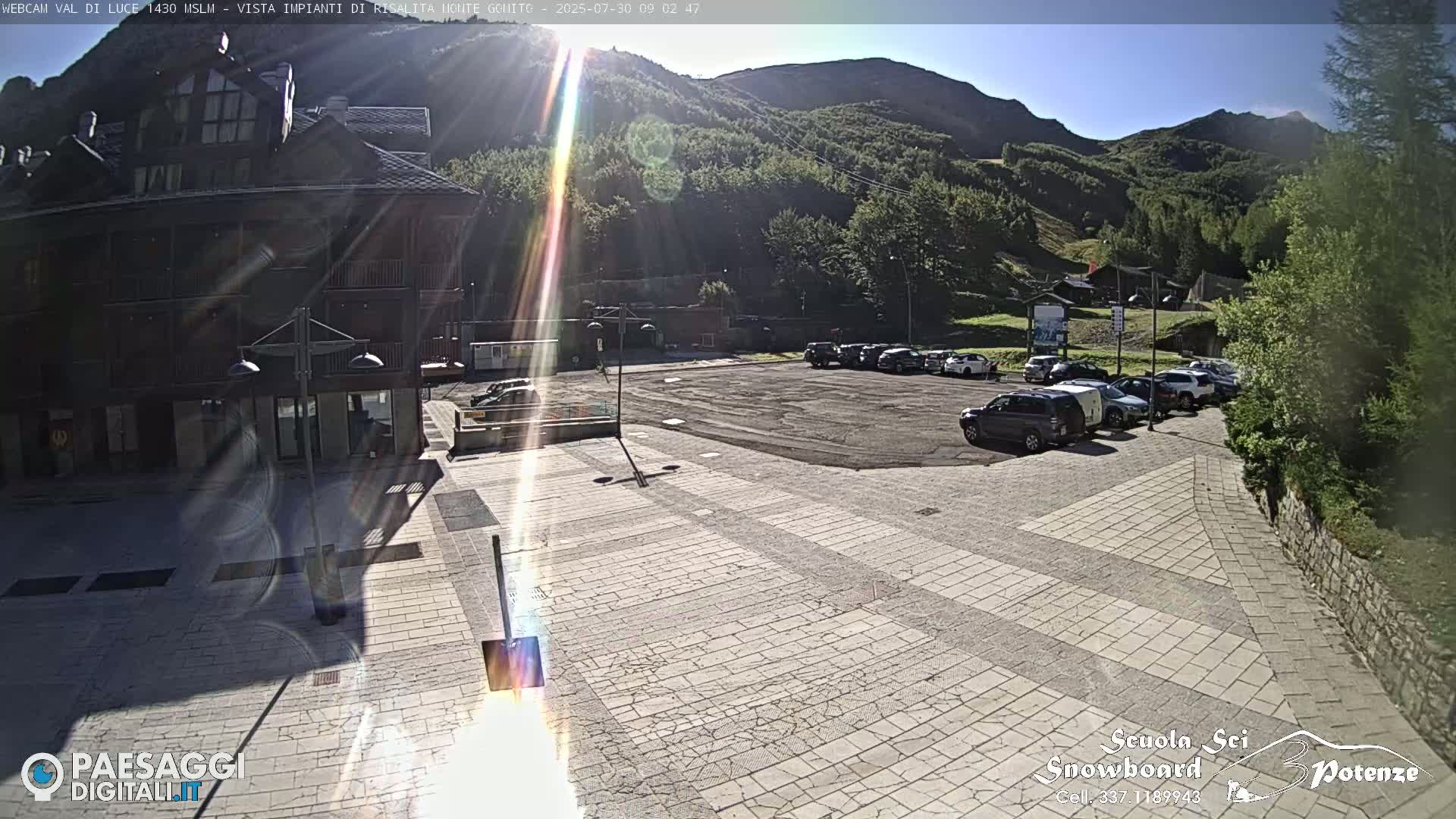 A sunny, paved plaza with a parking lot and a multi-story building is situated in front of a mountain range covered in lush green vegetation.