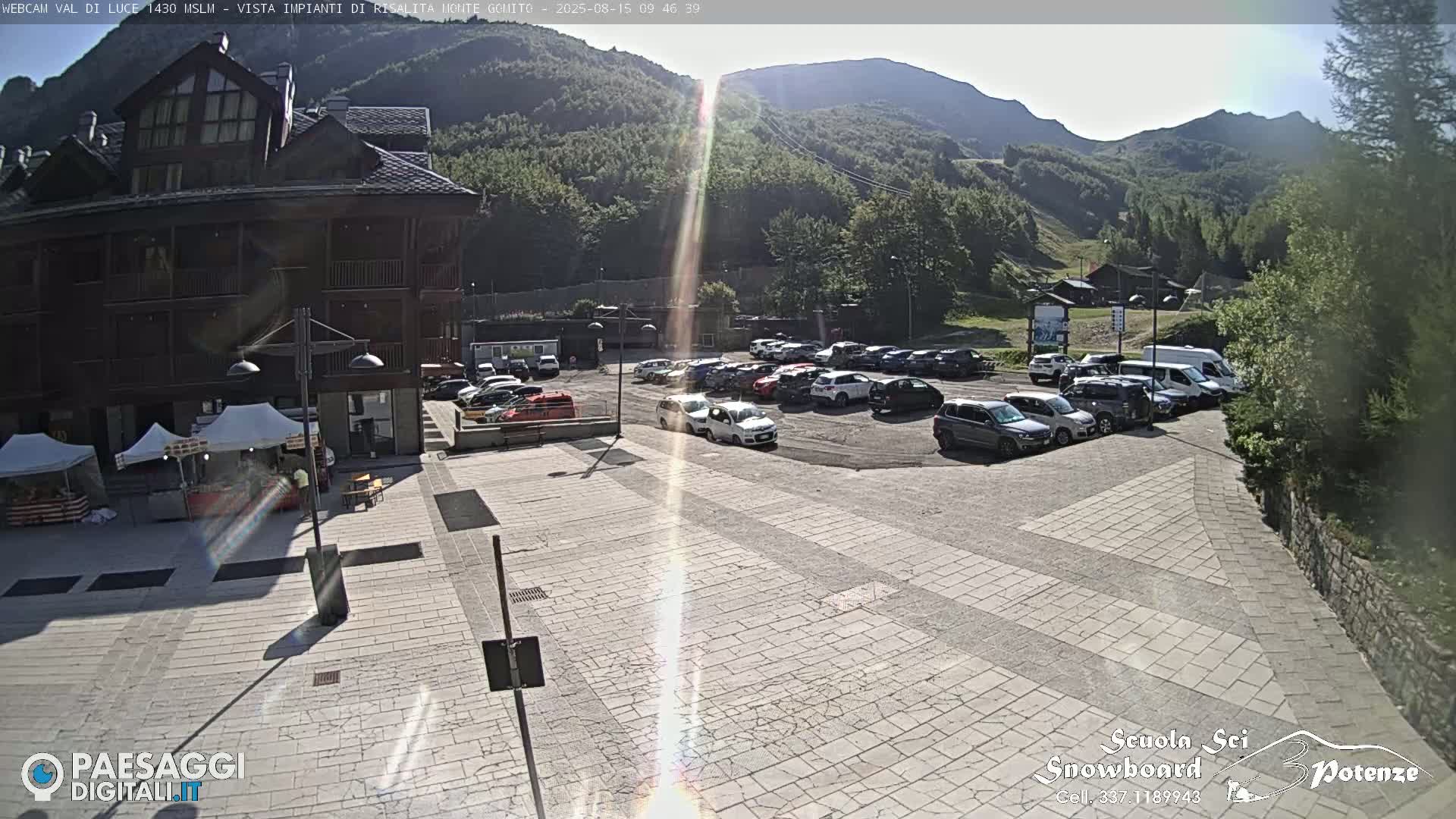 A sunny day reveals a paved plaza with a full parking lot and a large building, all nestled in a mountainous region.