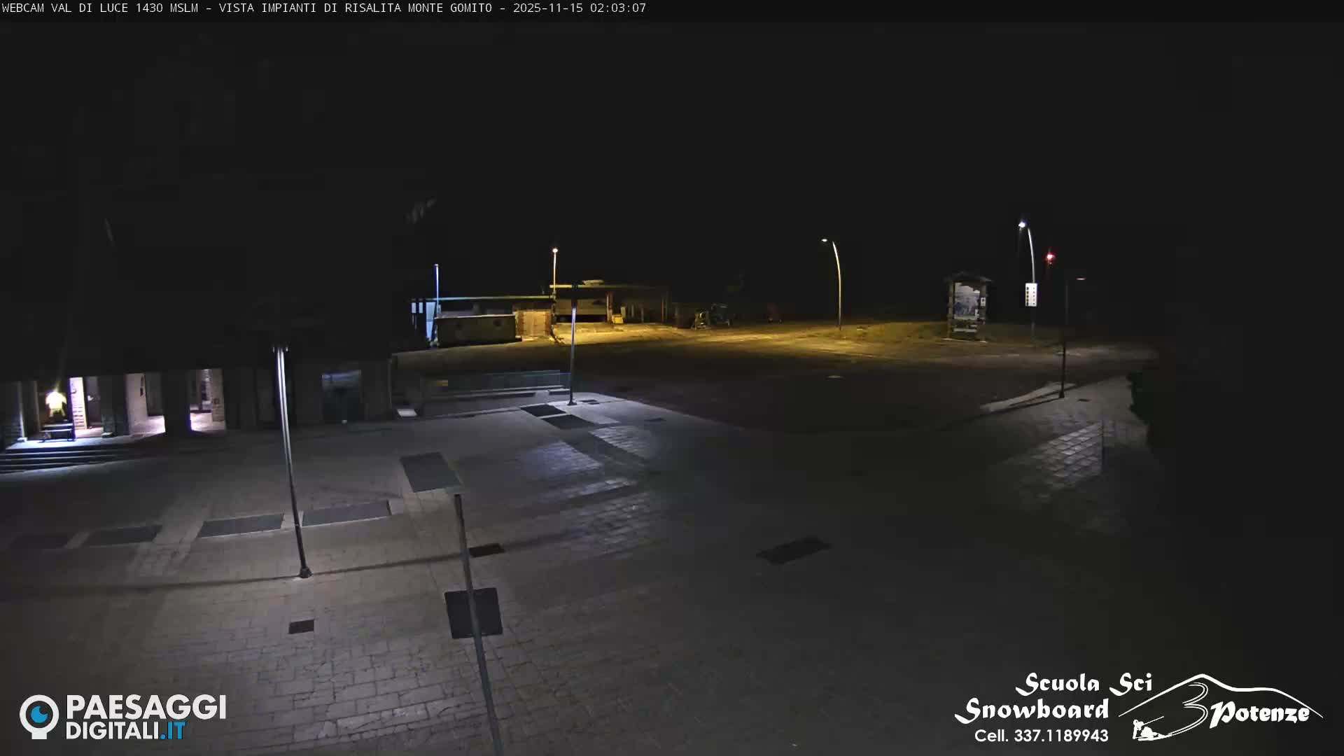 A deserted outdoor area is illuminated by streetlights at night, revealing paved surfaces, distant buildings, and a red traffic light under clear skies.